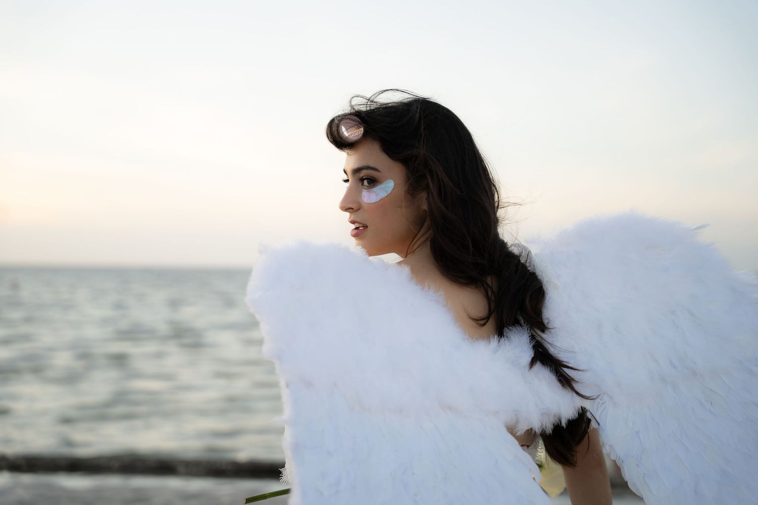 Young woman with angel wings and artistic makeup poses by ocean at dusk in white costume.