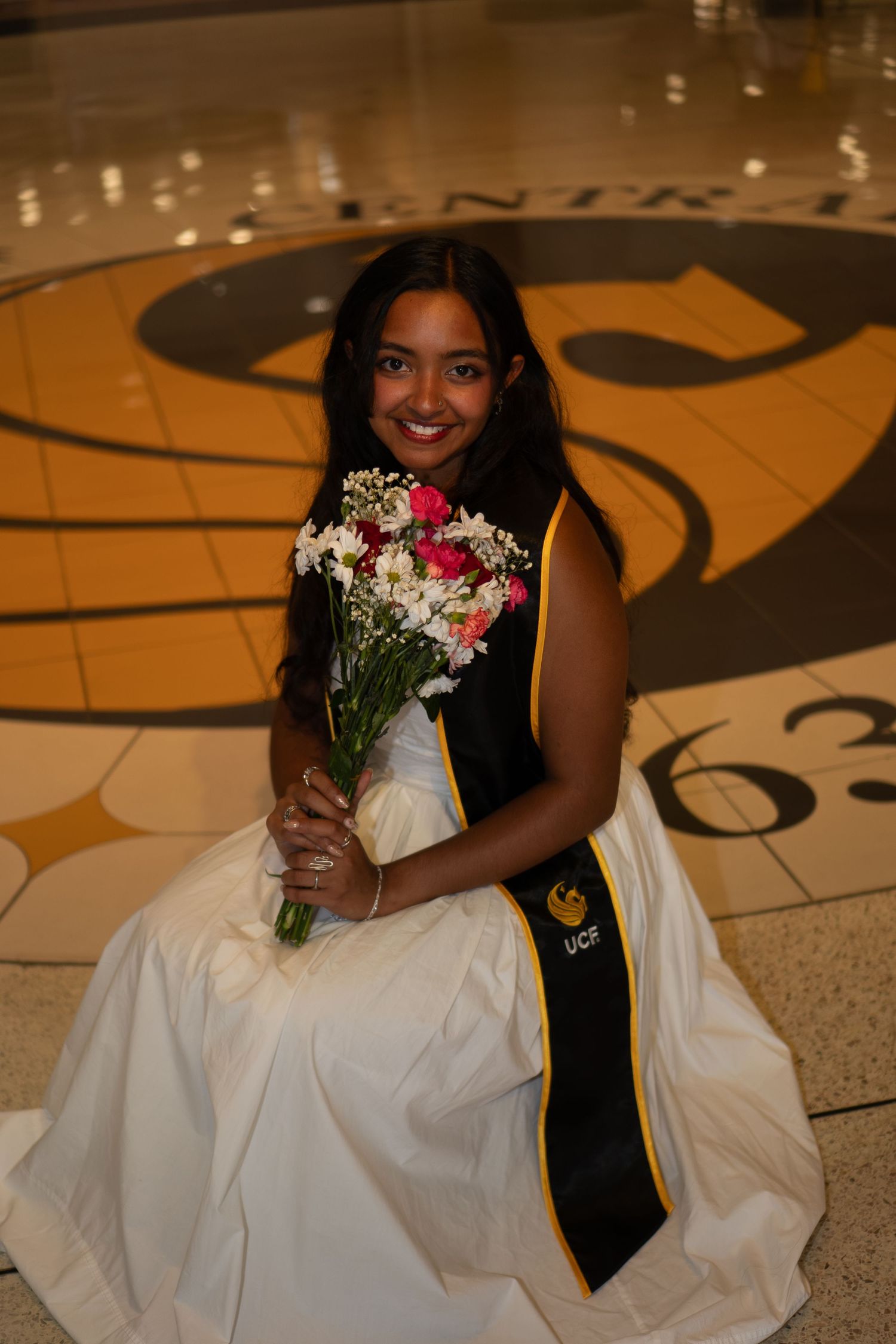 Smiling graduate in white dress holds pink flowers while kneeling on UCF logo floor.