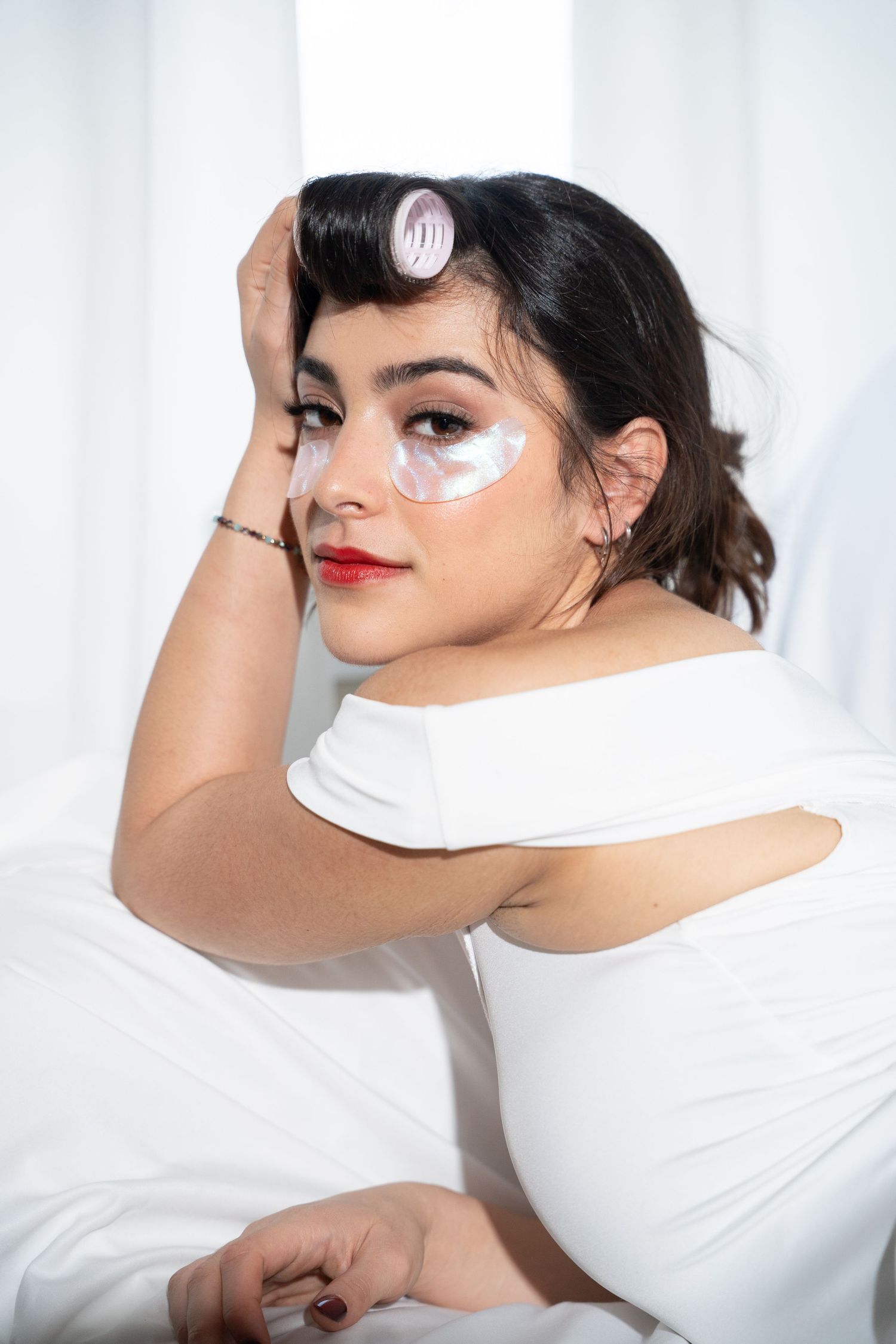 Woman in white off-shoulder dress with eye patches and hair roller posing against white background.
