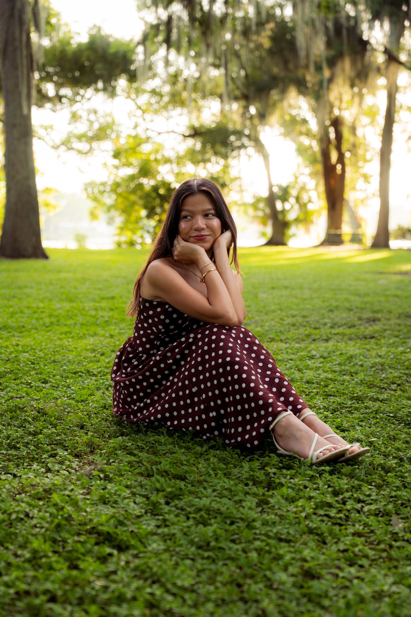 Young woman in burgundy polka dot dress sitting on green grass in sunlit park with hands on chin.