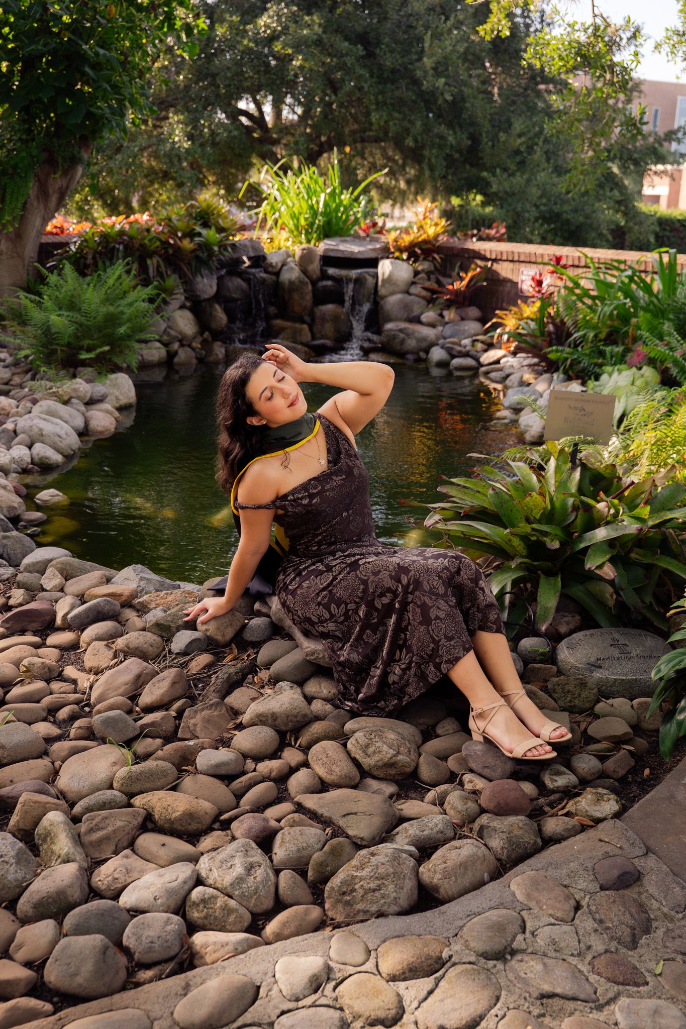 Woman in brown floral dress poses on river rocks beside a serene garden pond with waterfall.