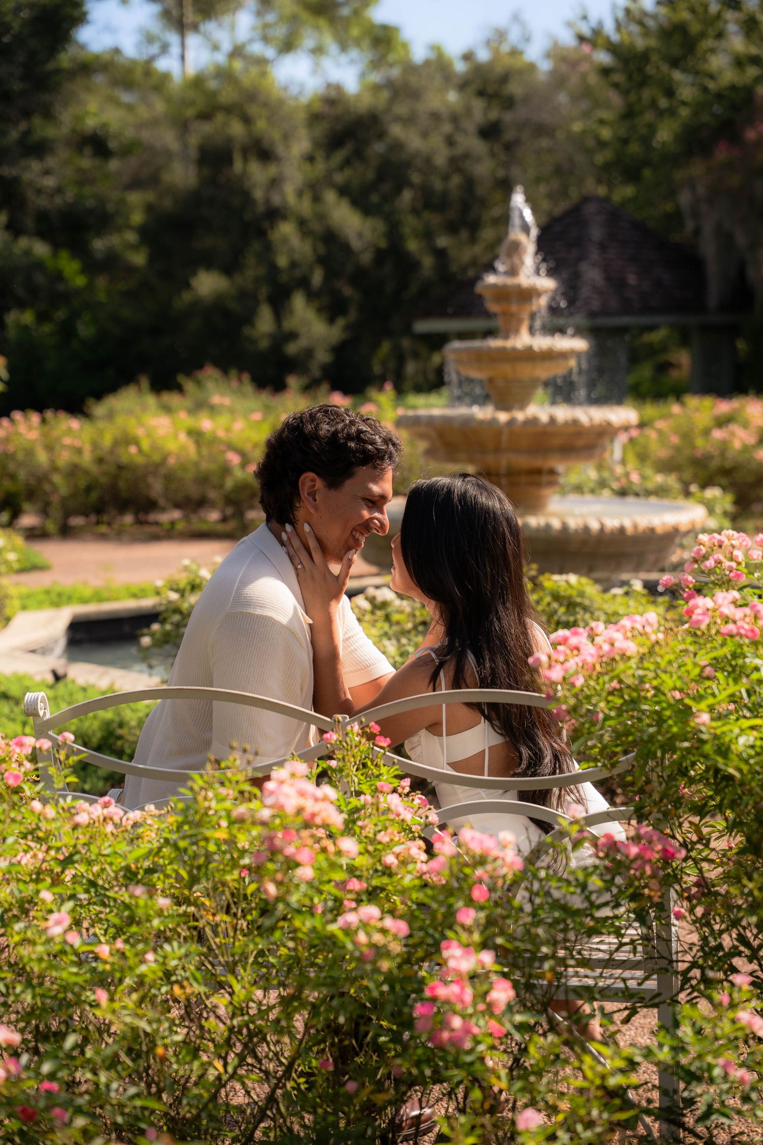 Romantic couple sitting close together on bench surrounded by pink roses near garden fountain.