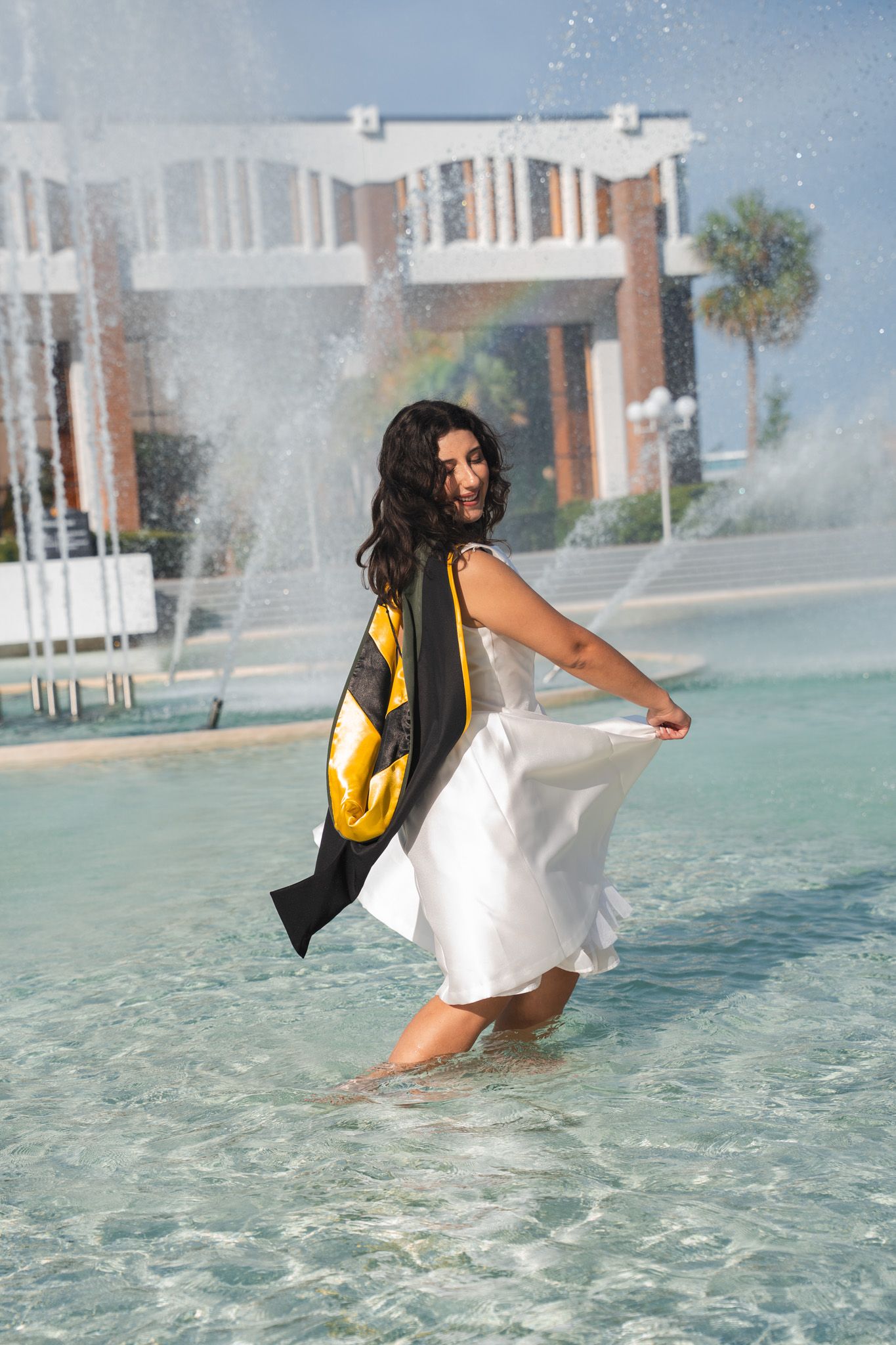 Smiling graduate in white dress wades through fountain water wearing black and gold academic hood.