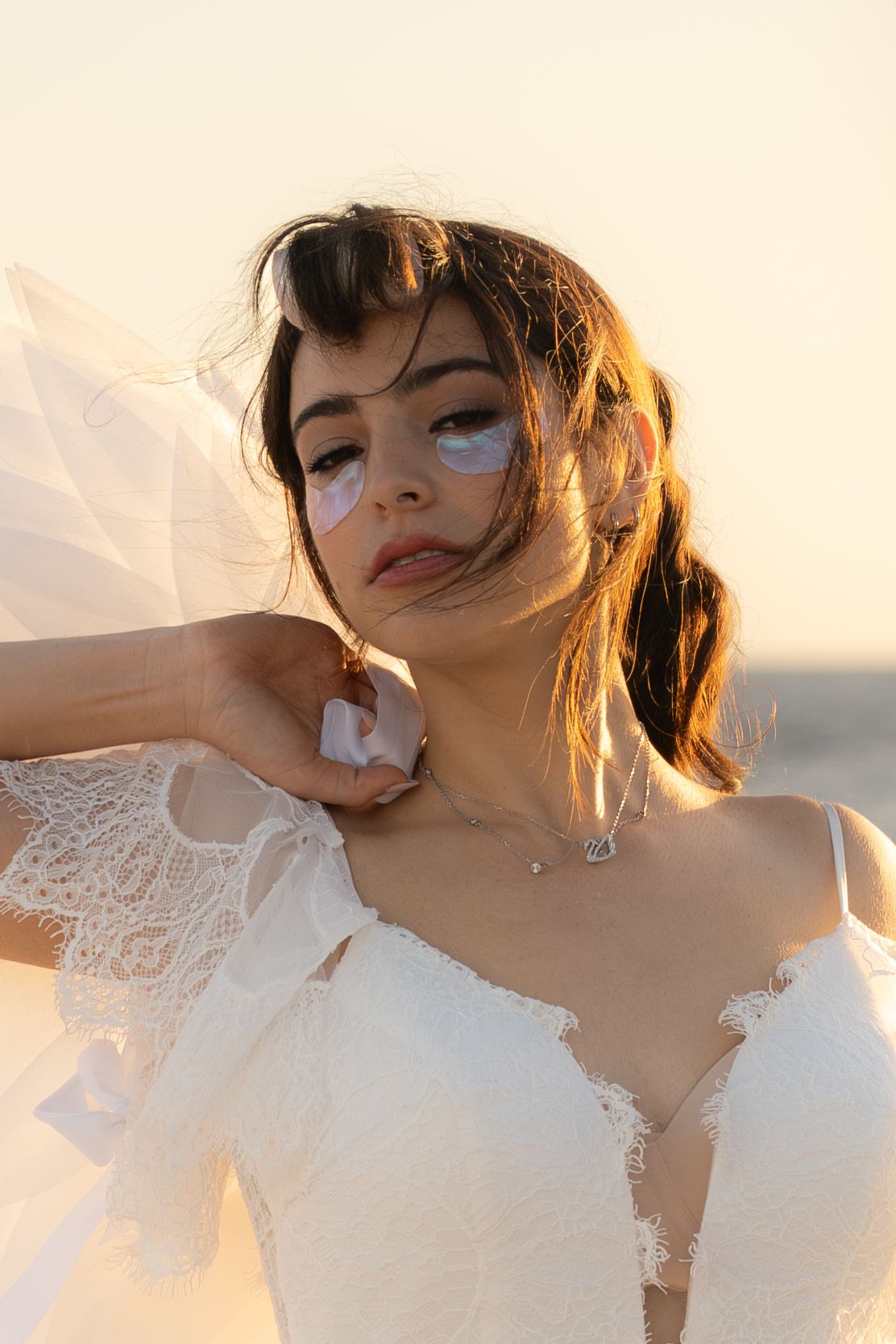 Woman in white lace bridal gown with eye patches poses in golden sunset light outdoors.