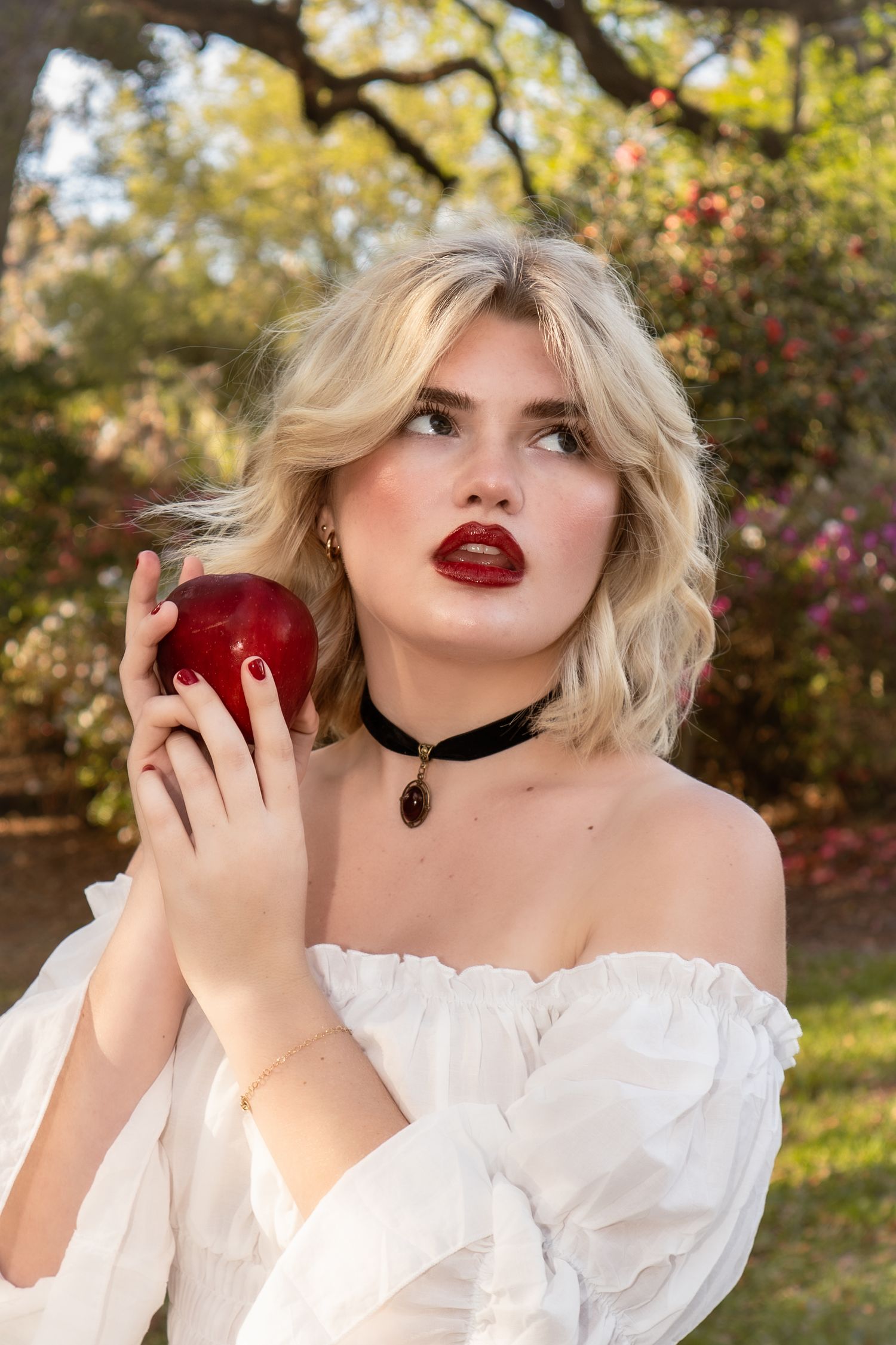 Blonde woman in white off-shoulder top holding red apple outdoors in a garden setting.