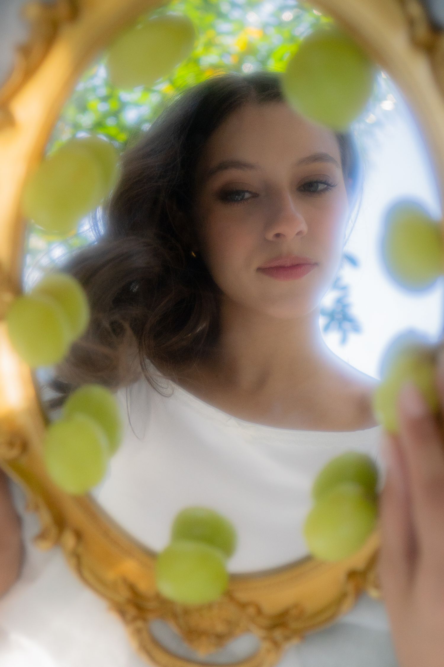 Brunette woman reflected in ornate gold mirror decorated with green apples outdoors.