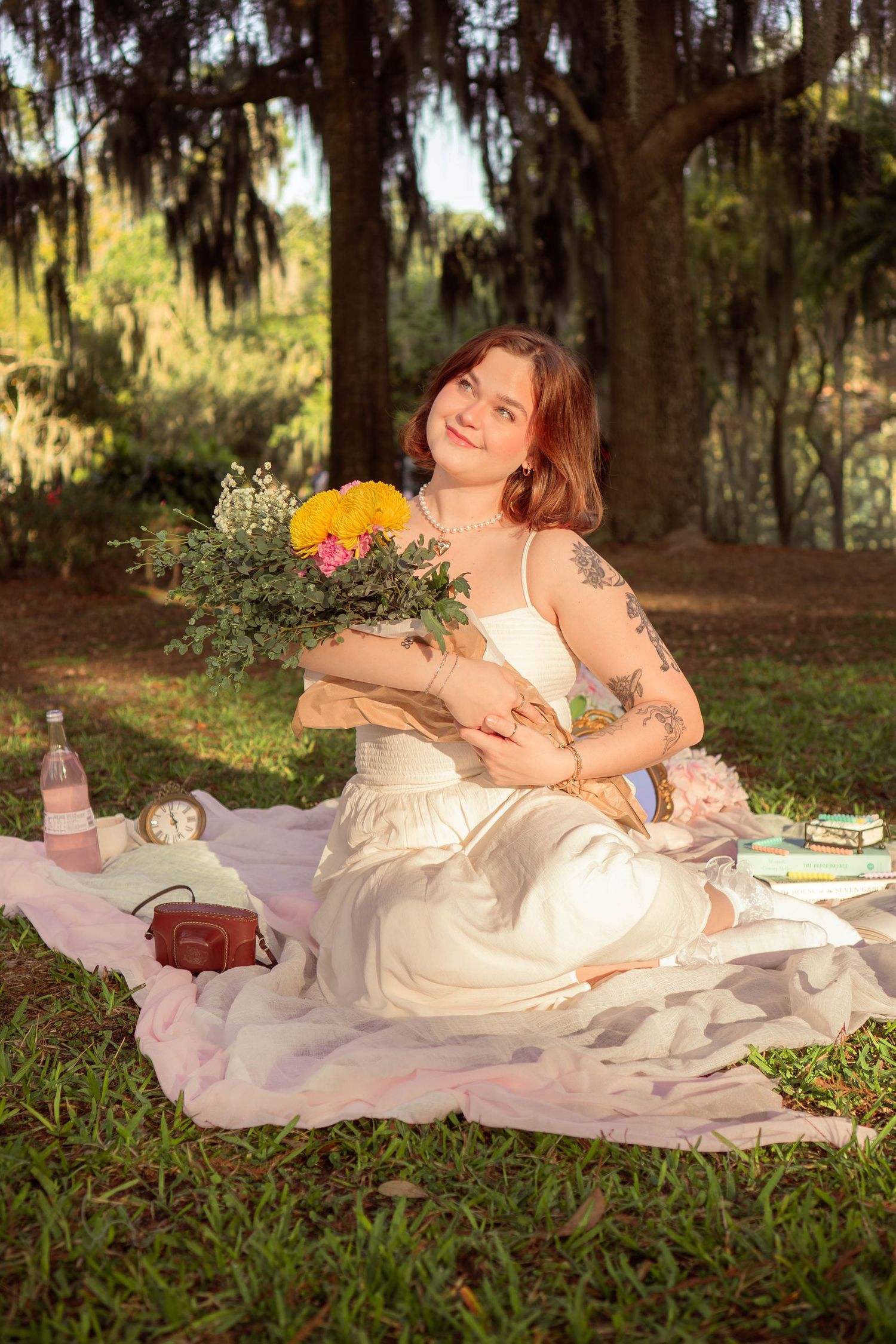 Young woman in white dress sitting on blanket in park holding colorful flower bouquet.