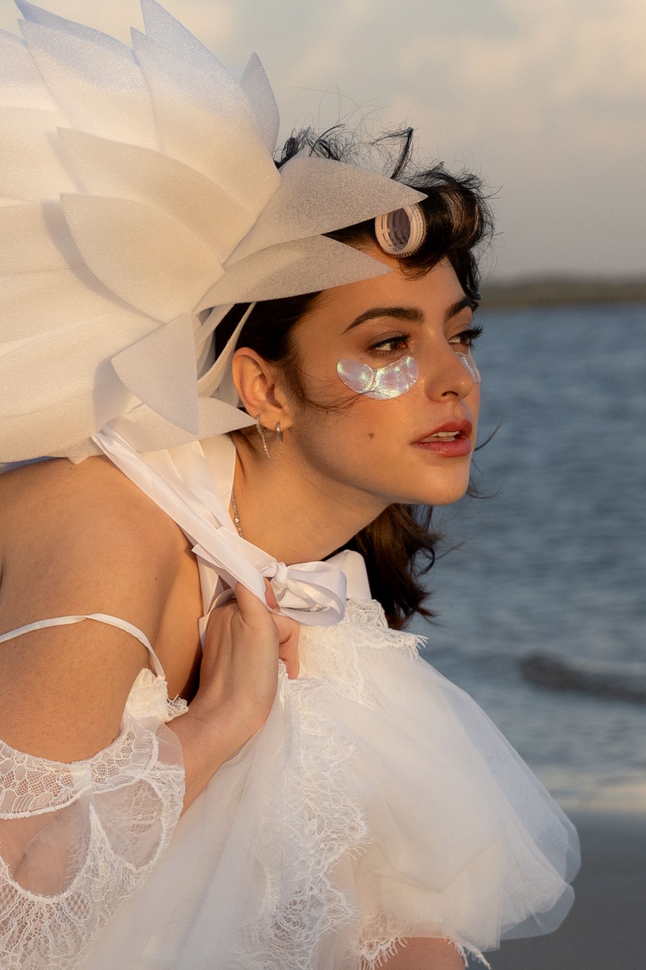 Close-up of woman in white lace outfit with feathered headpiece and artistic eye makeup by the sea.