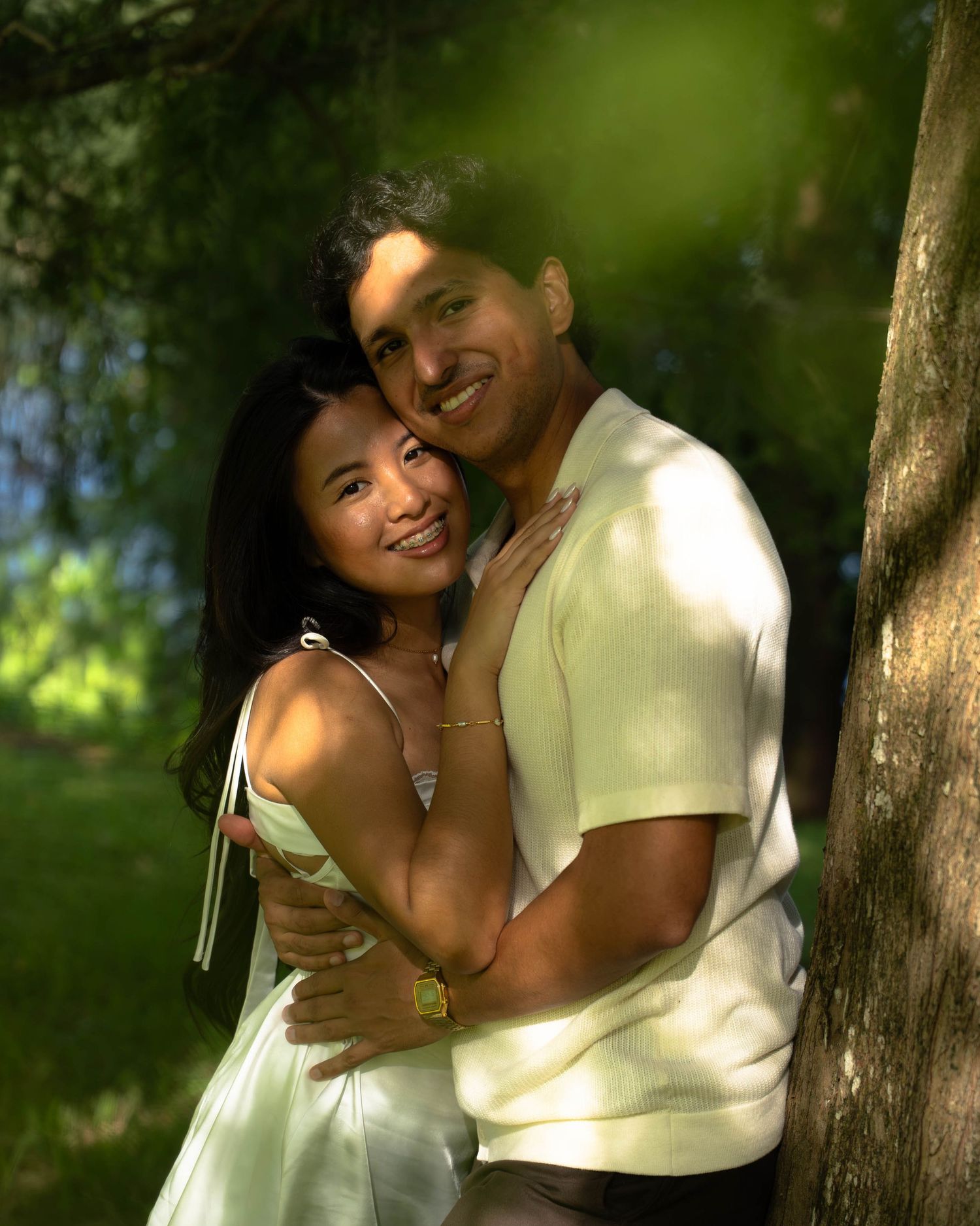 A happy couple embraces near a tree in a sunny outdoor engagement photo session.