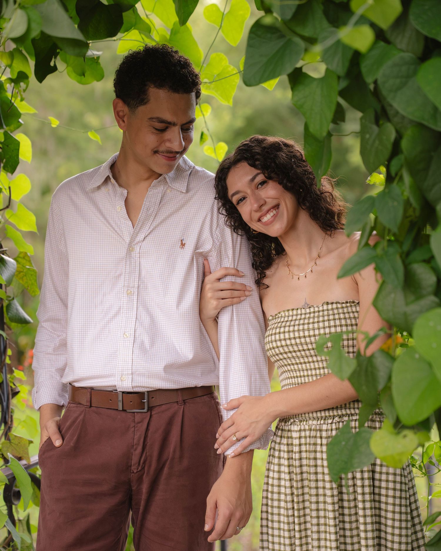A young couple poses romantically under a lush green vine archway outdoors.