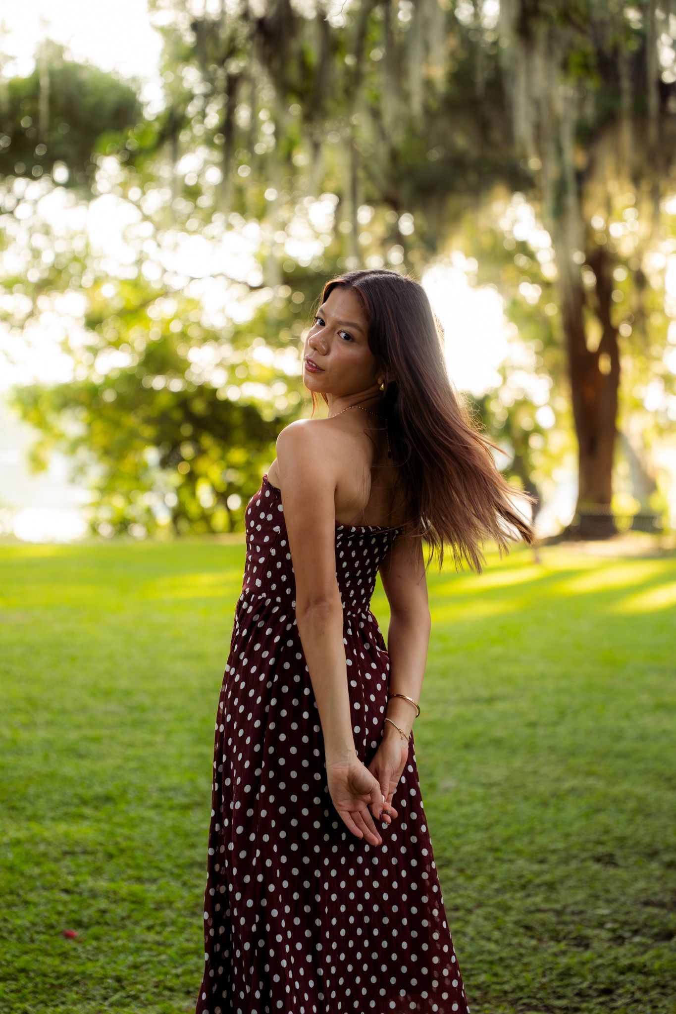 Young woman in strapless burgundy polka dot dress standing on lush green lawn with hair blowing in breeze.