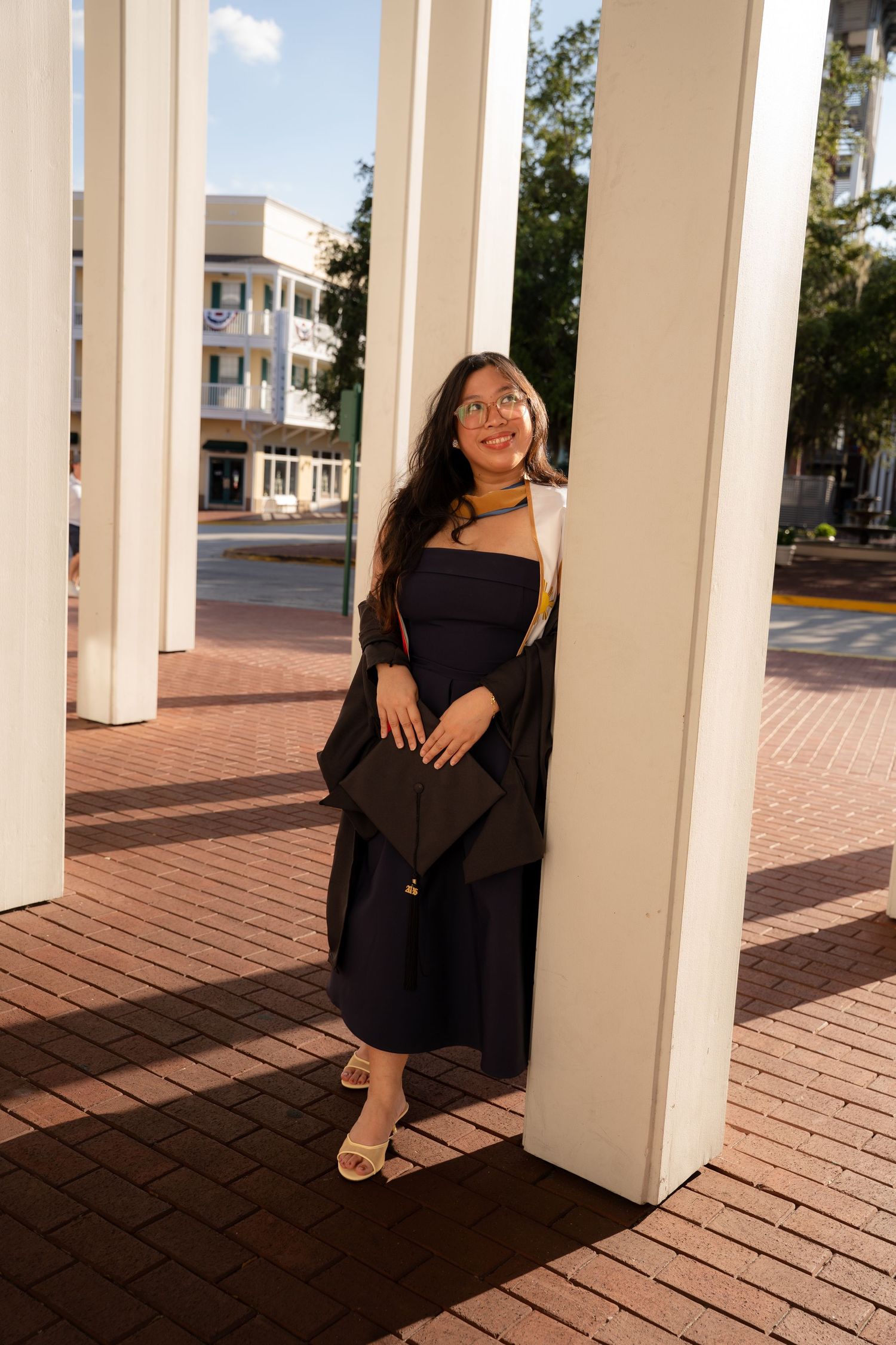 Young woman in black graduation gown and gold stole poses between white columns on brick plaza.