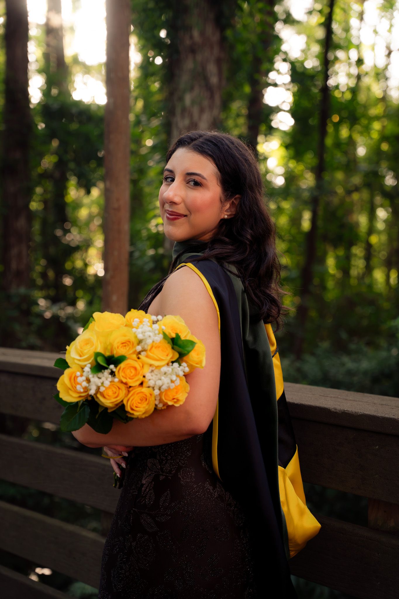 Graduate in black gown holds yellow rose bouquet on wooden bridge in forest setting.