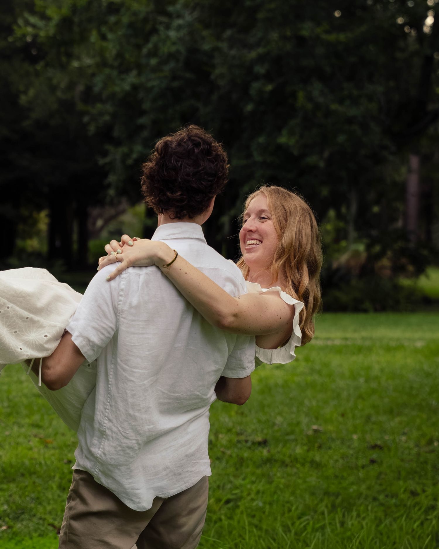 Man joyfully lifting and spinning a laughing woman in a white dress on green grass.