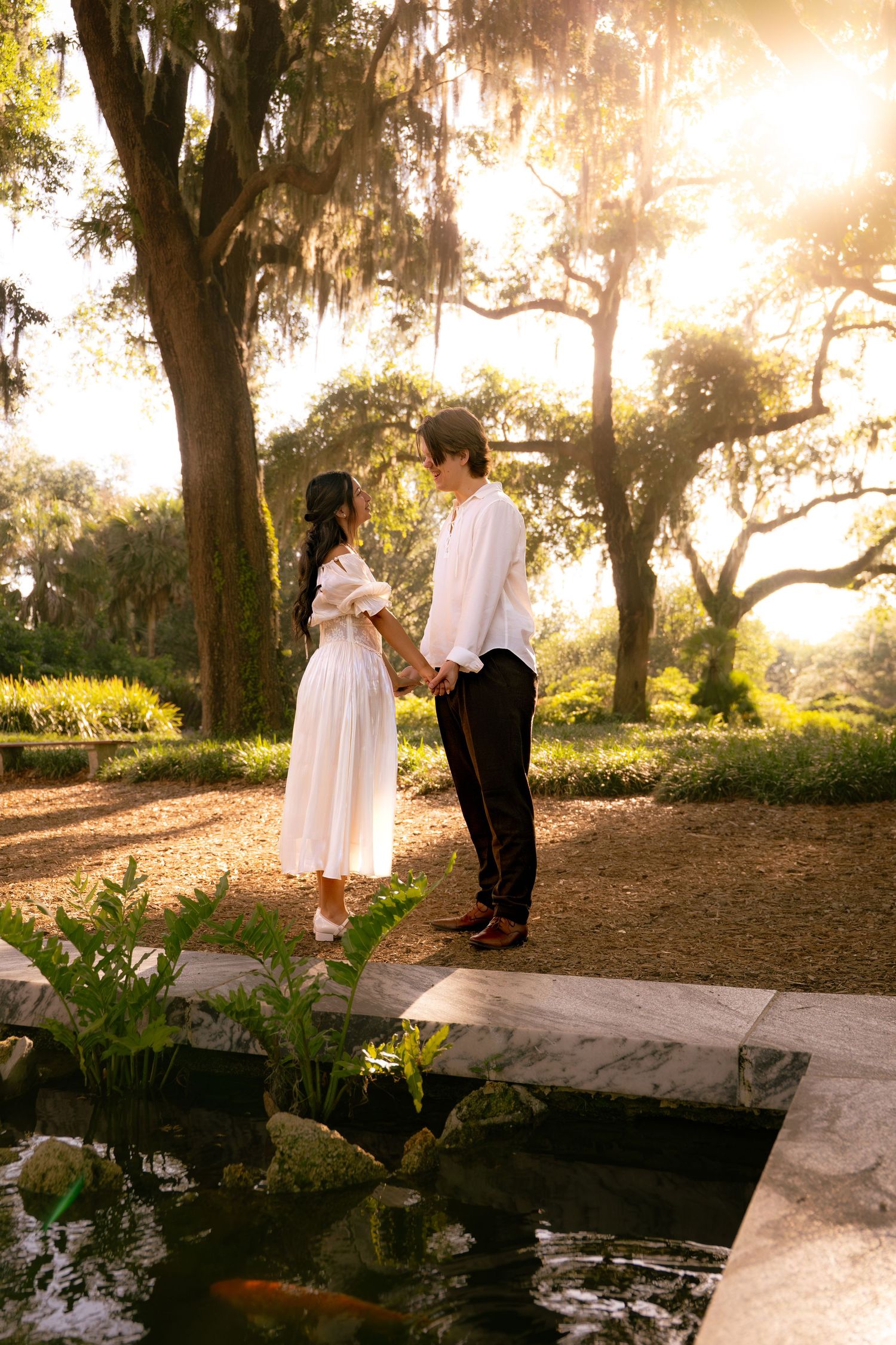 Couple holding hands by a koi pond in a sunlit garden with Spanish moss-draped oak trees.