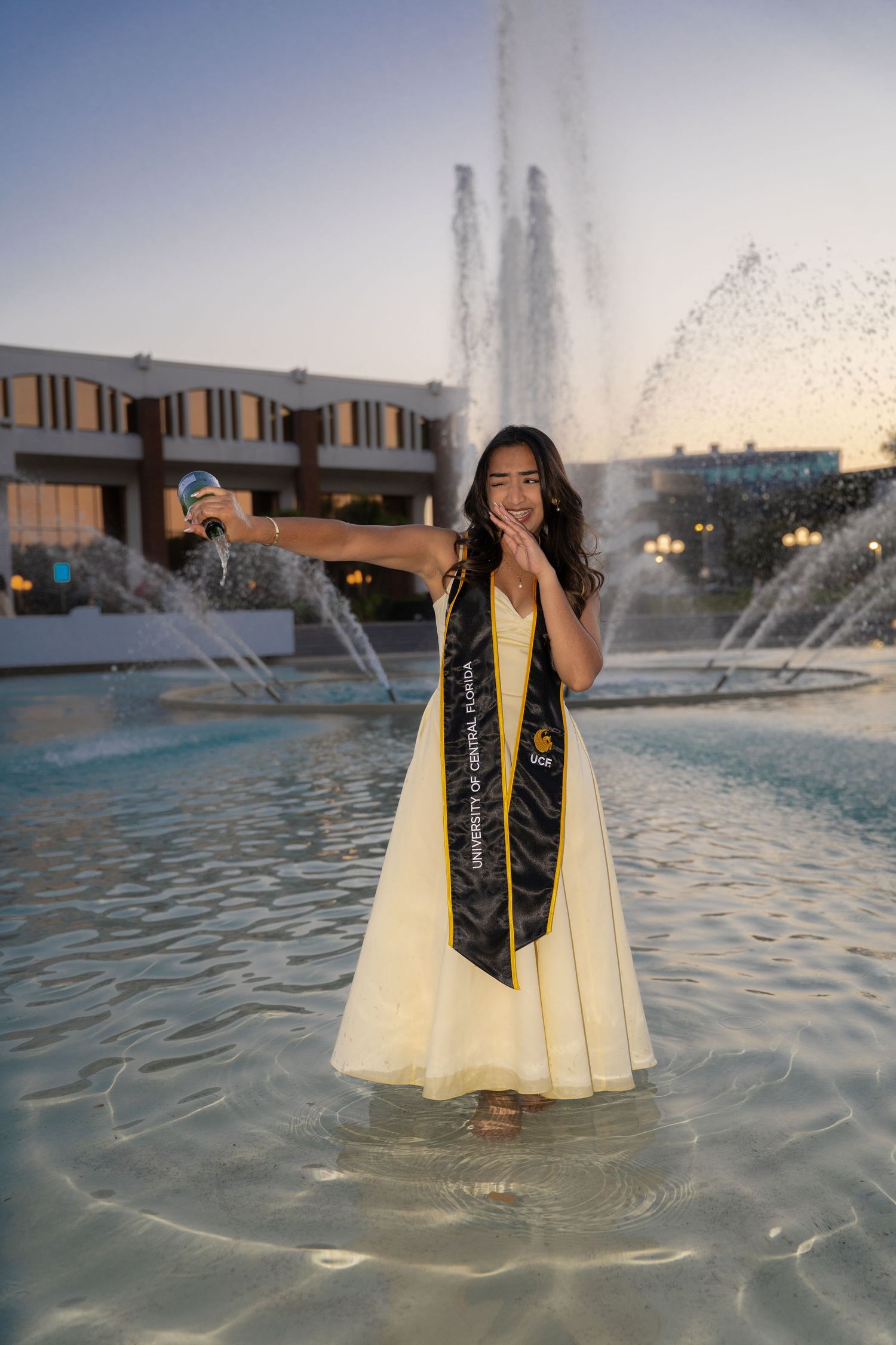 UCF graduate in yellow dress stands in campus fountain wearing black graduation stole at sunset.
