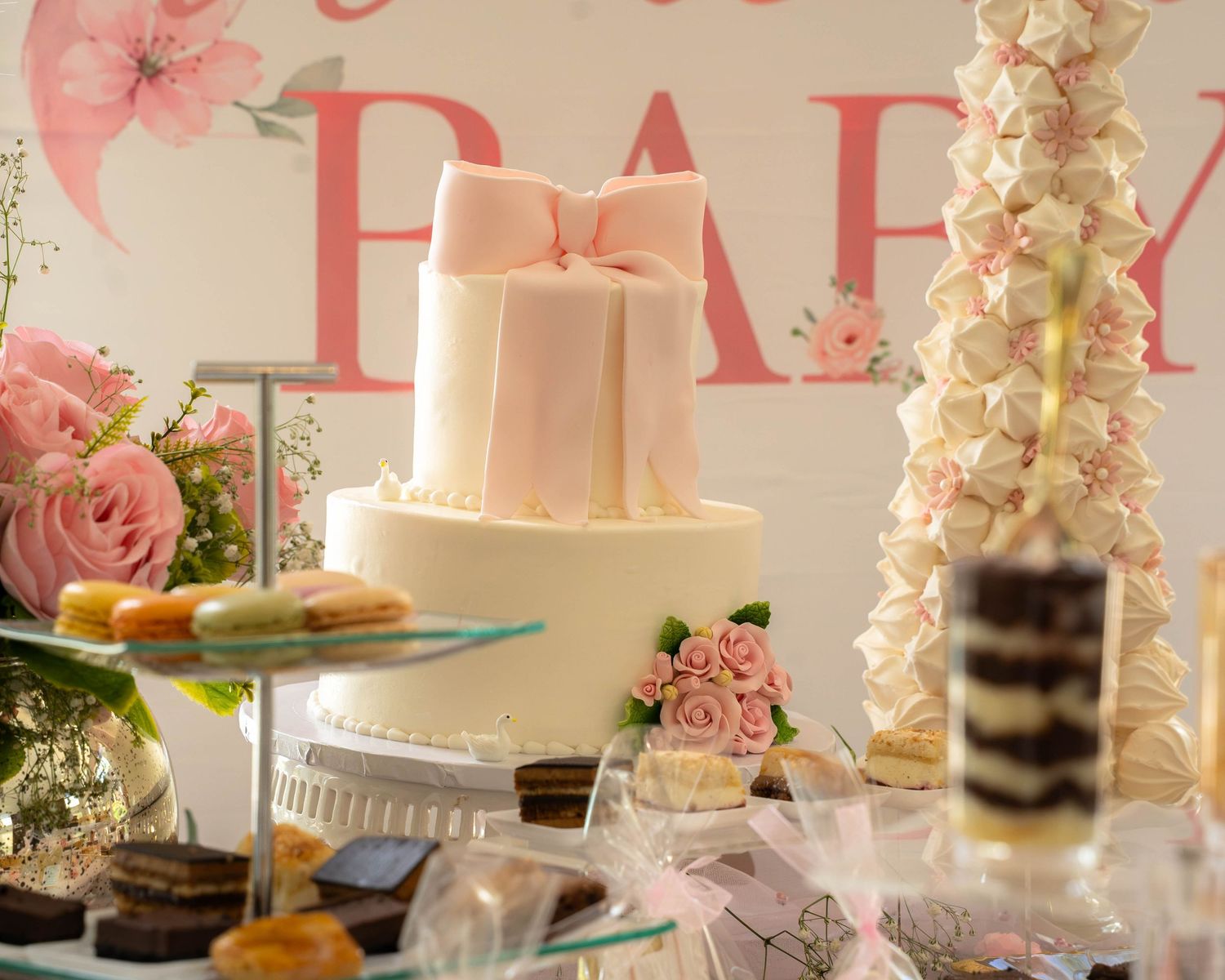 Elegant two-tier white baby shower cake with pink bow and roses beside a macaron tower and desserts.