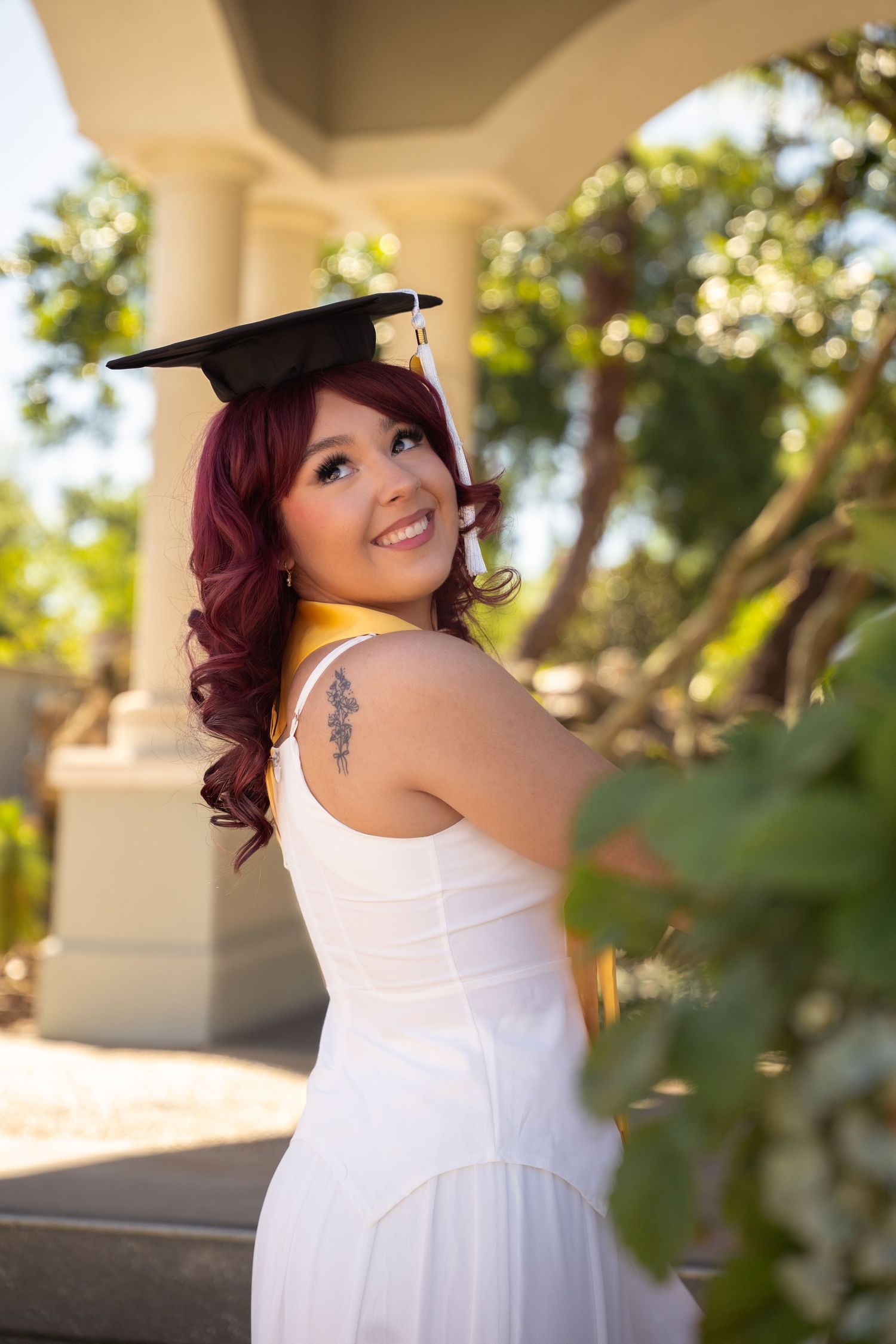 Smiling red-haired graduate in white dress and cap looks back over shoulder in garden setting.