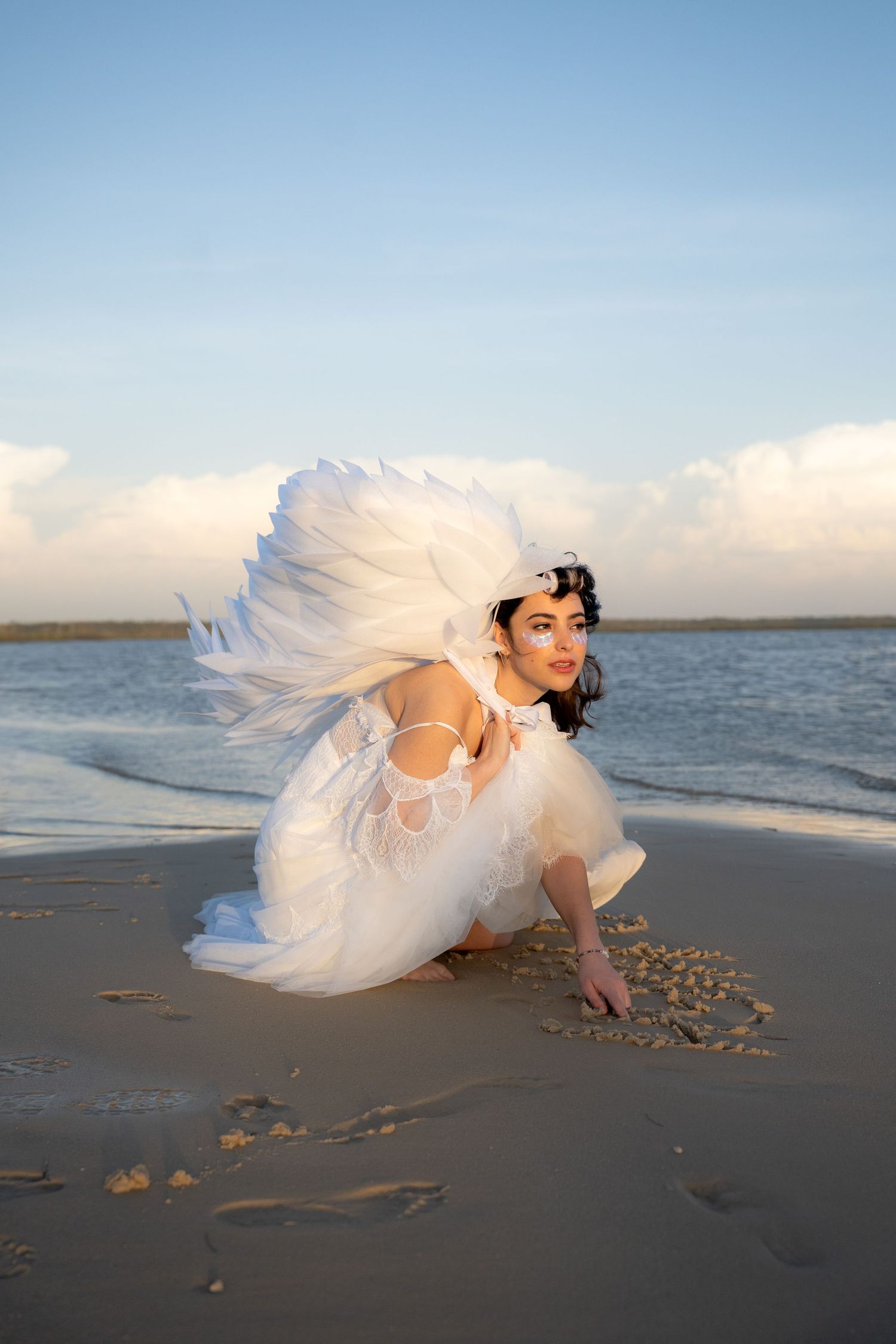 Woman in white angel costume with large wings crouching on sandy beach by the ocean.