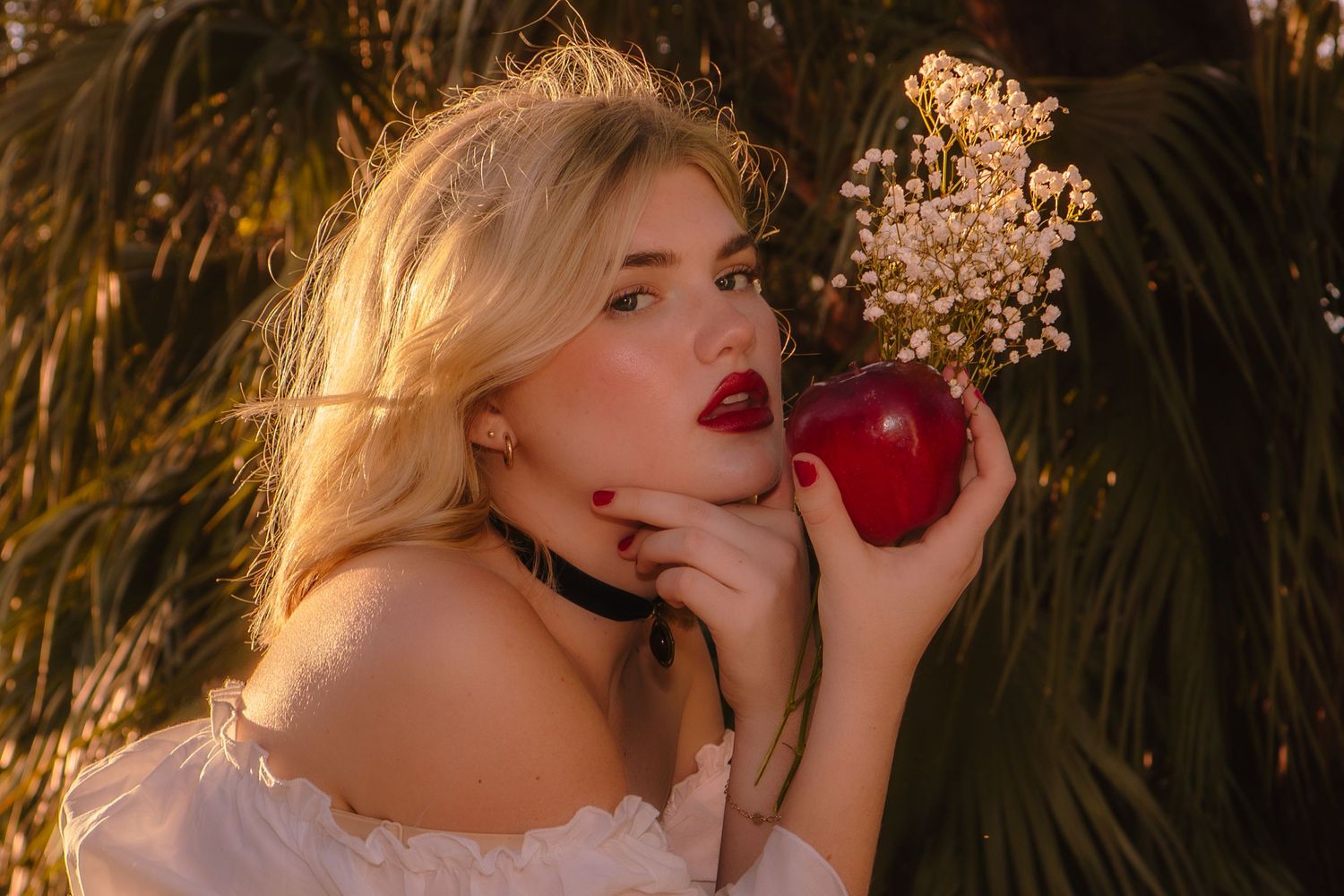 Blonde woman with red lips holding red apple and white flowers near tropical foliage.