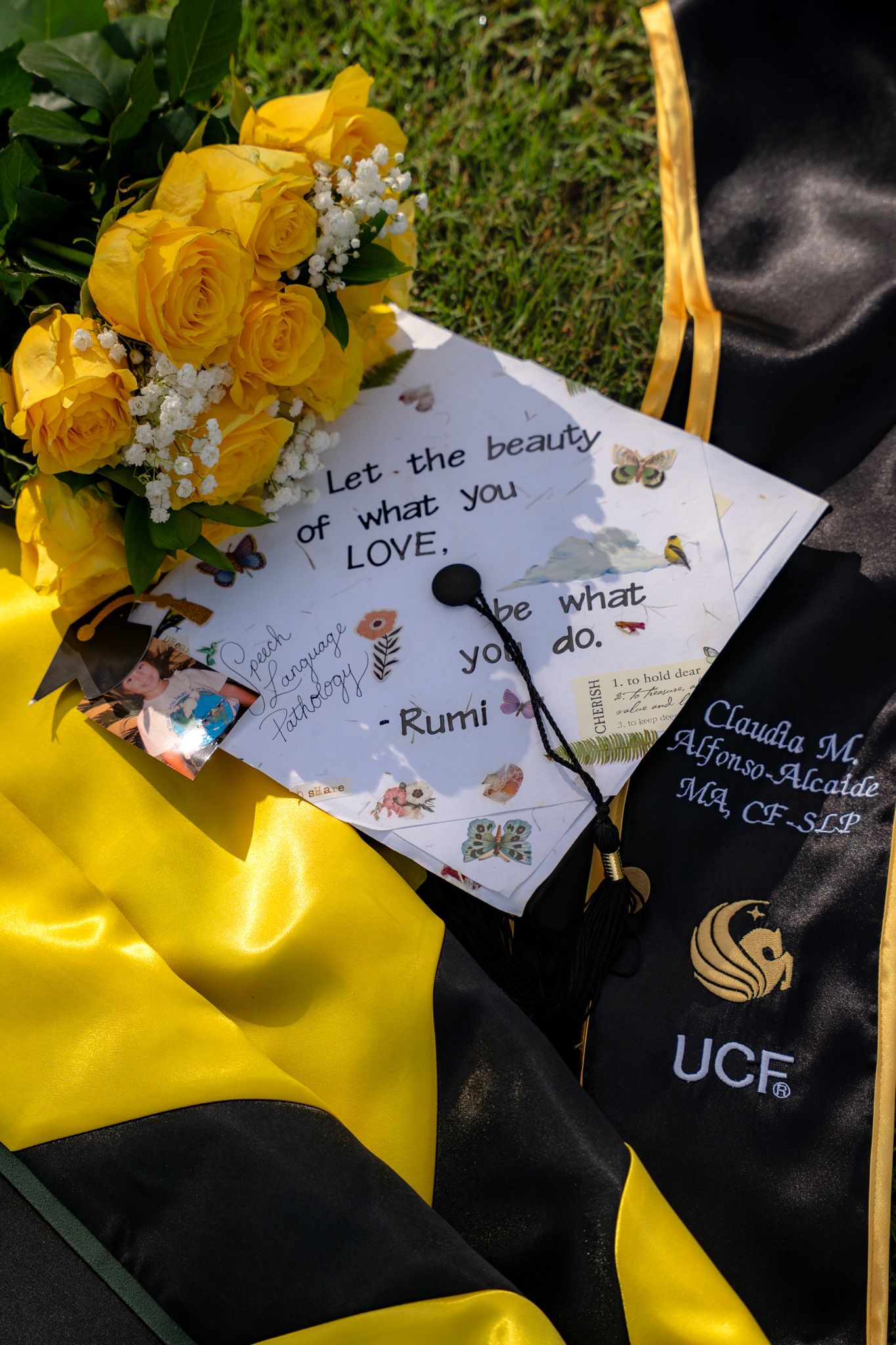 UCF graduation cap with Rumi quote beside yellow roses and black gold commencement regalia on grass.