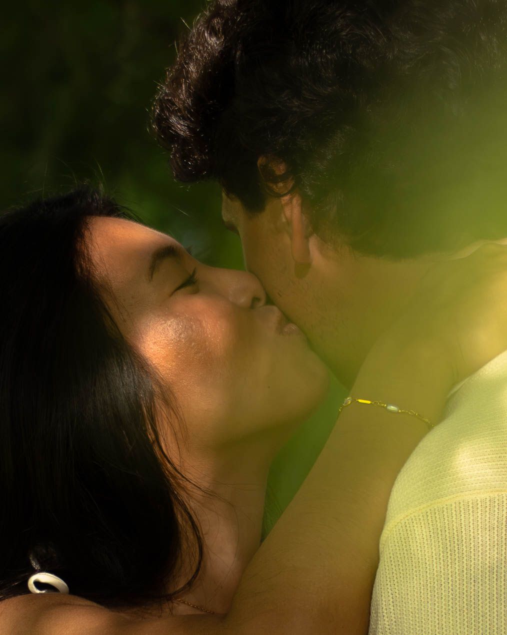 A couple shares a tender kiss outdoors in a lush green garden setting.