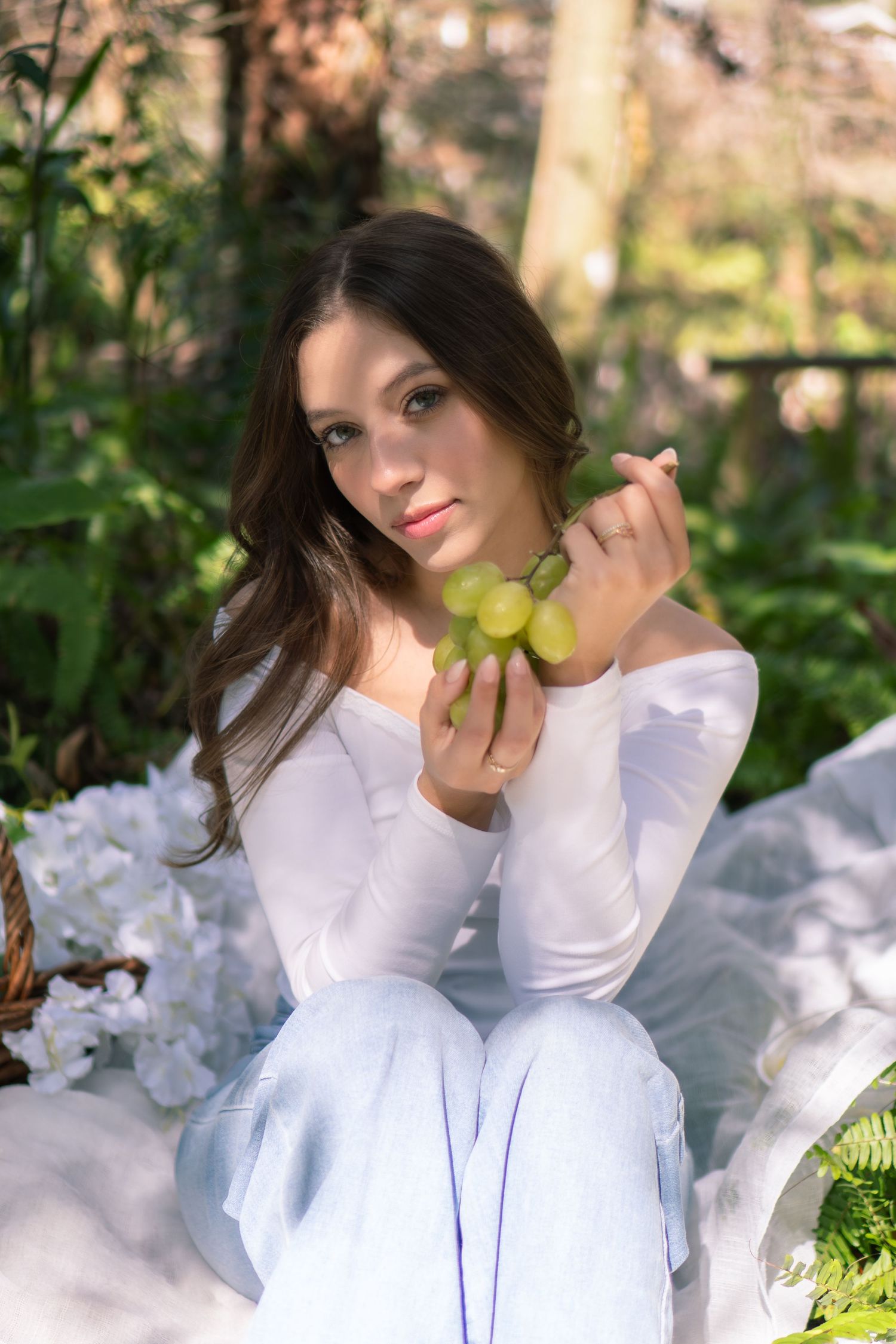 A young brunette woman in white holds green grapes during an outdoor garden picnic setting.
