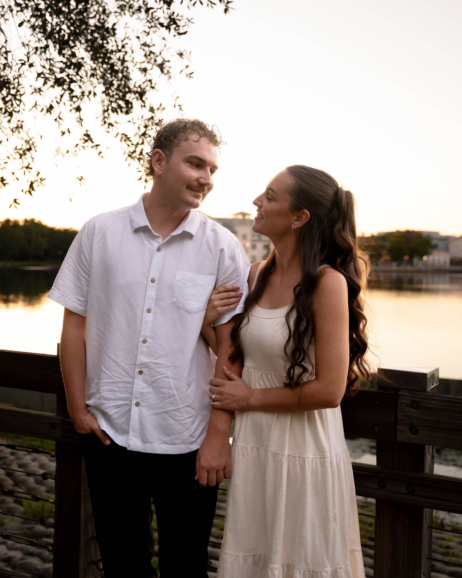 Couple in white outfits sharing a romantic moment on a wooden bridge at sunset near a serene lake.