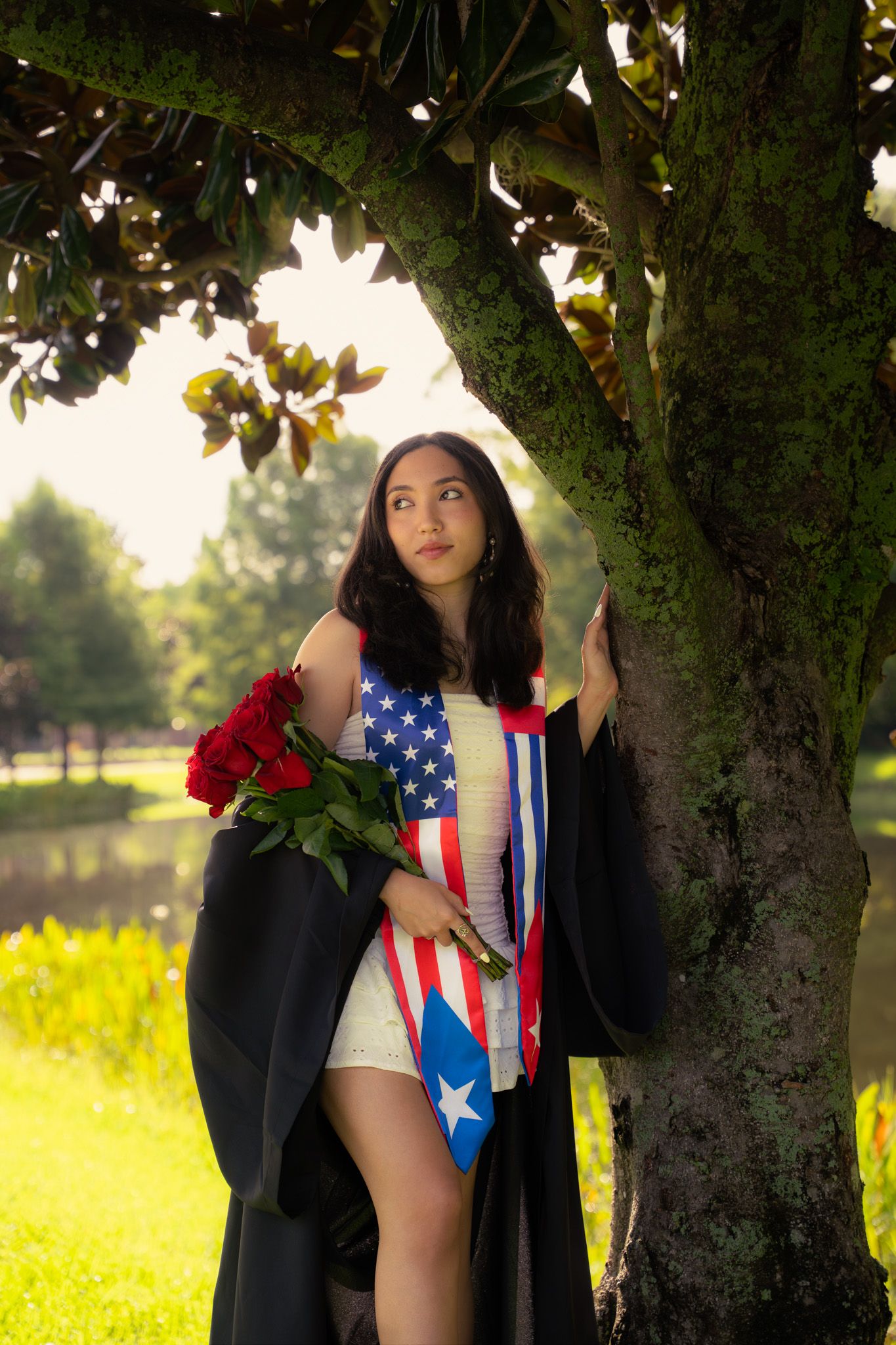 Female graduate in black gown with American and Puerto Rican flags holds red roses beside mossy tree.