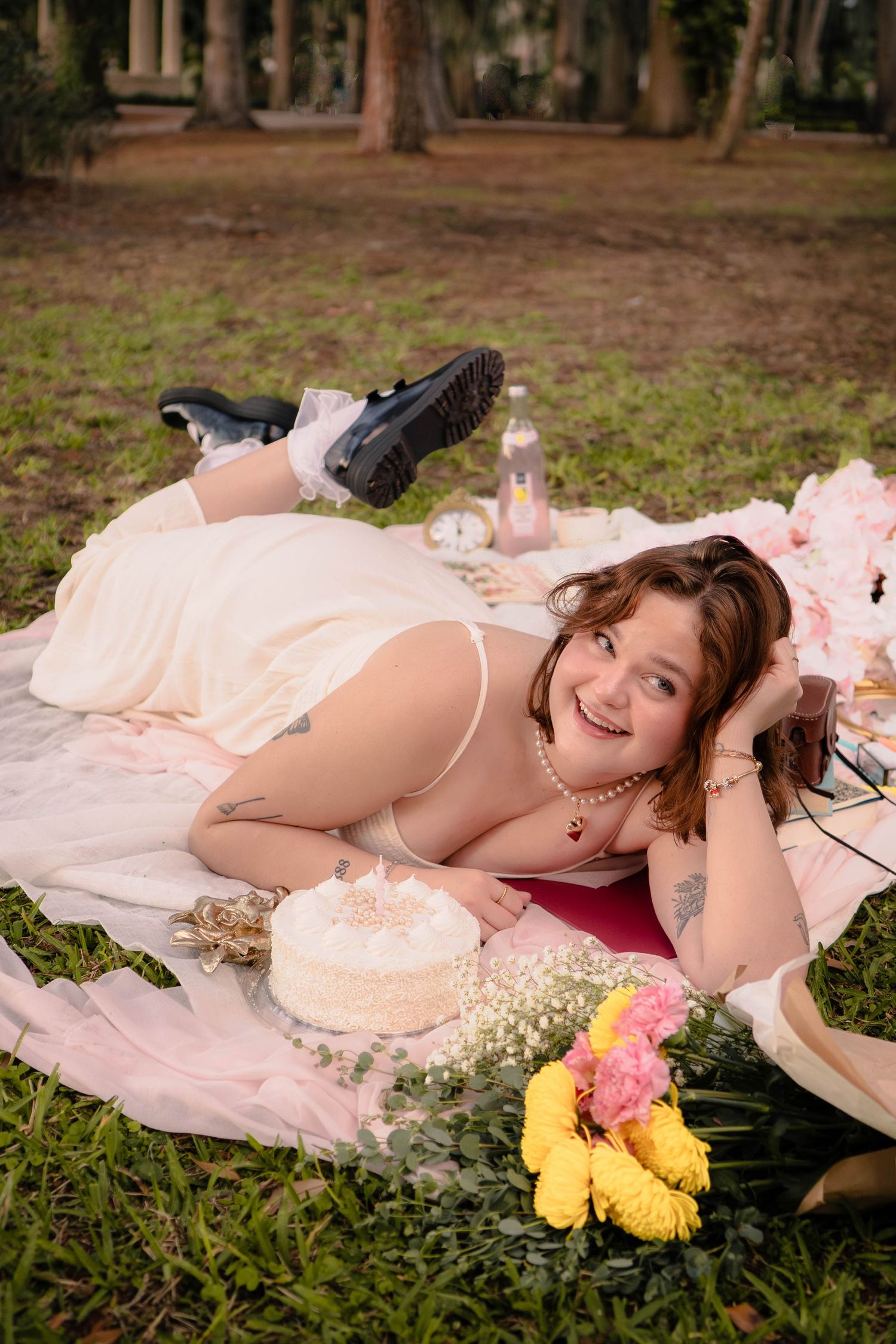 Smiling woman in lingerie lying on a picnic blanket with cake and colorful flowers in a park.
