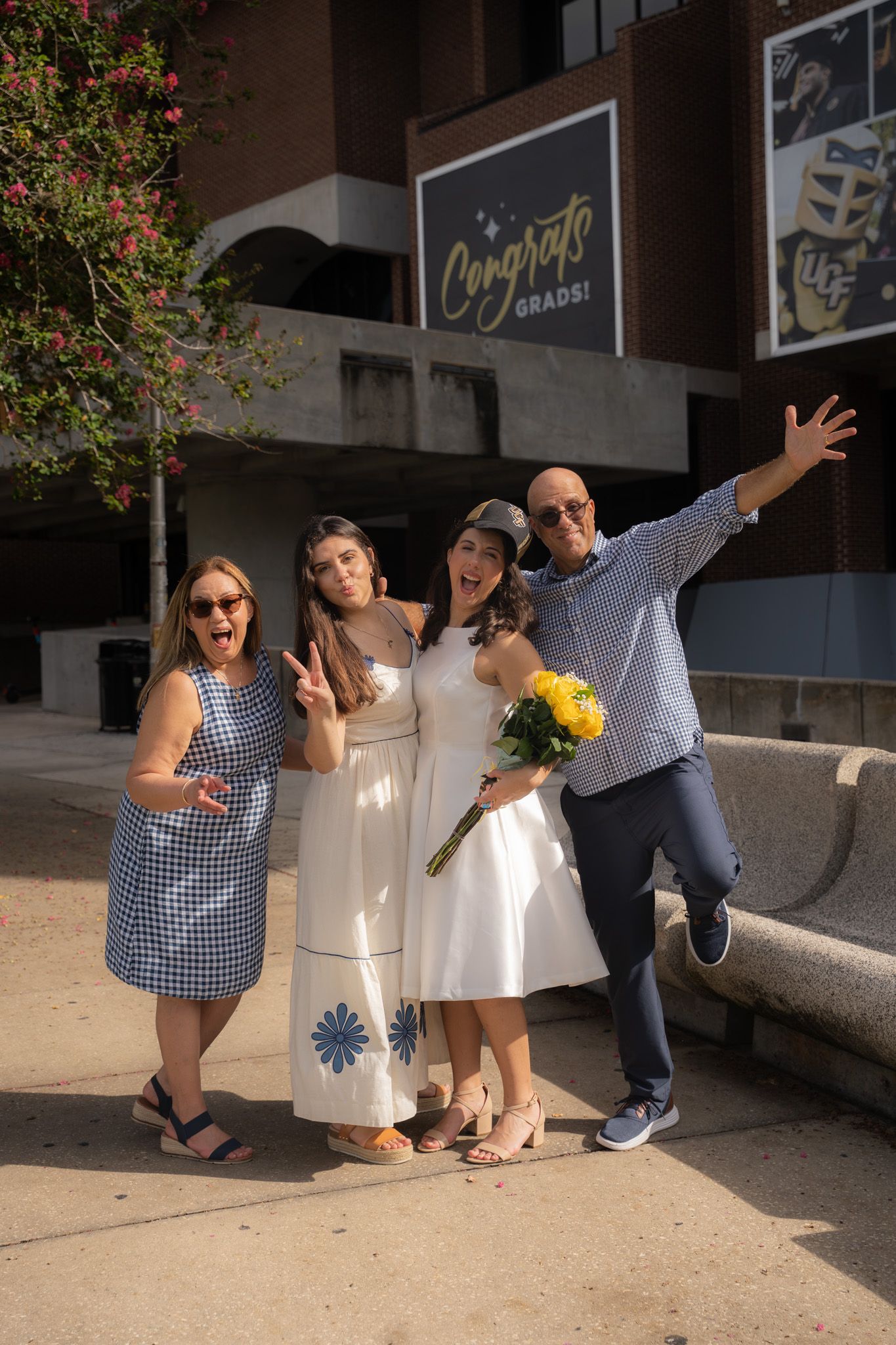 Happy family of four celebrates graduation outside venue with Congrats Grads sign and yellow roses.