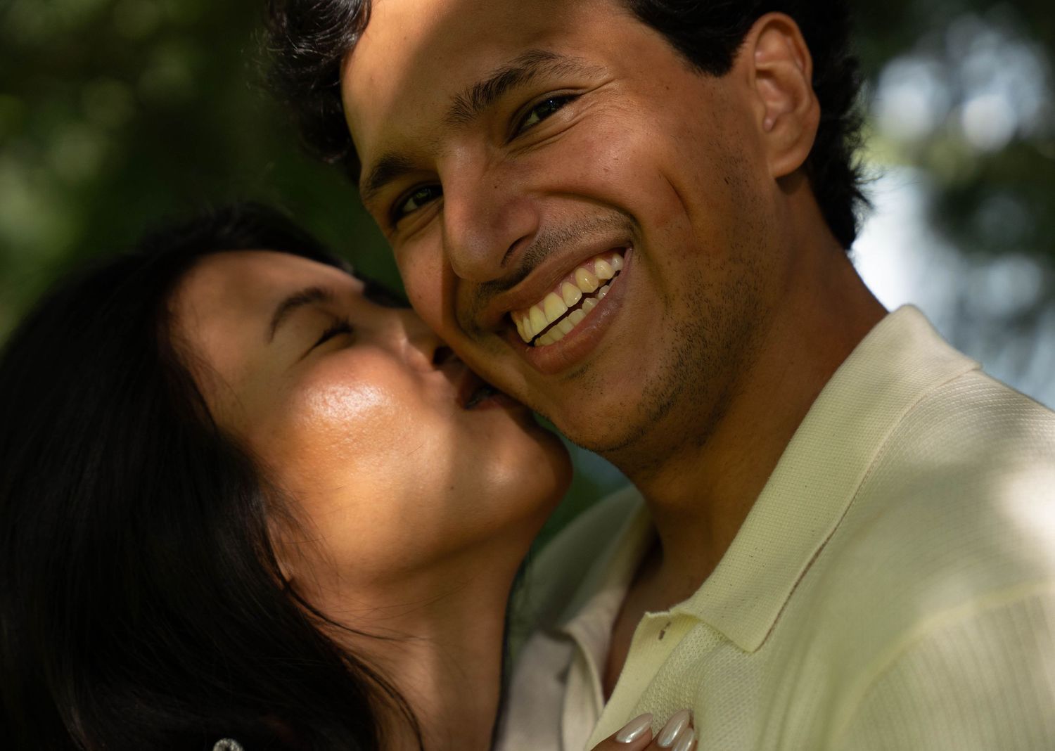 A woman kisses a smiling man on the cheek outdoors in a romantic engagement photo session.
