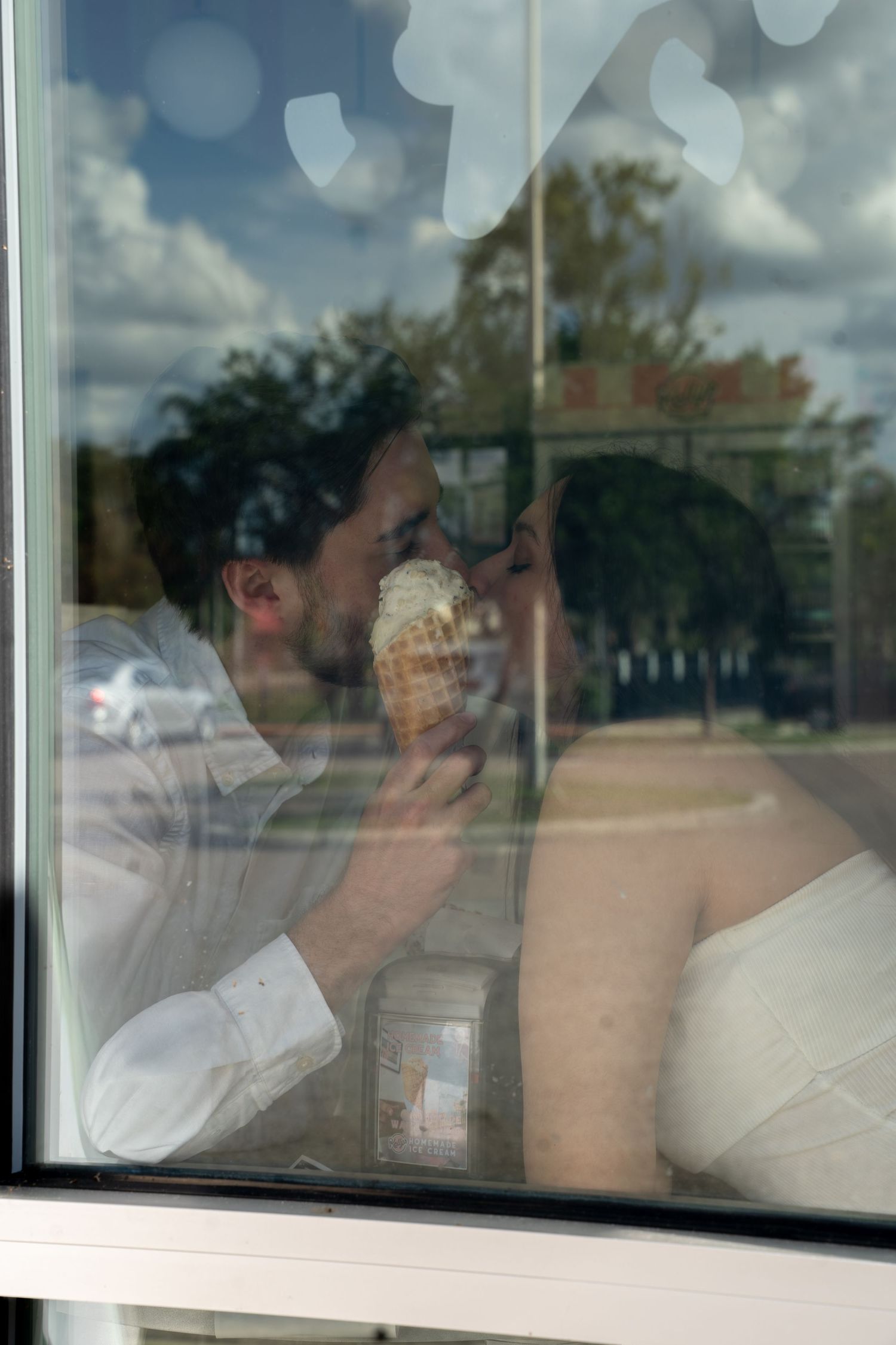 Bride and groom sharing an ice cream cone kiss reflected in a shop window.