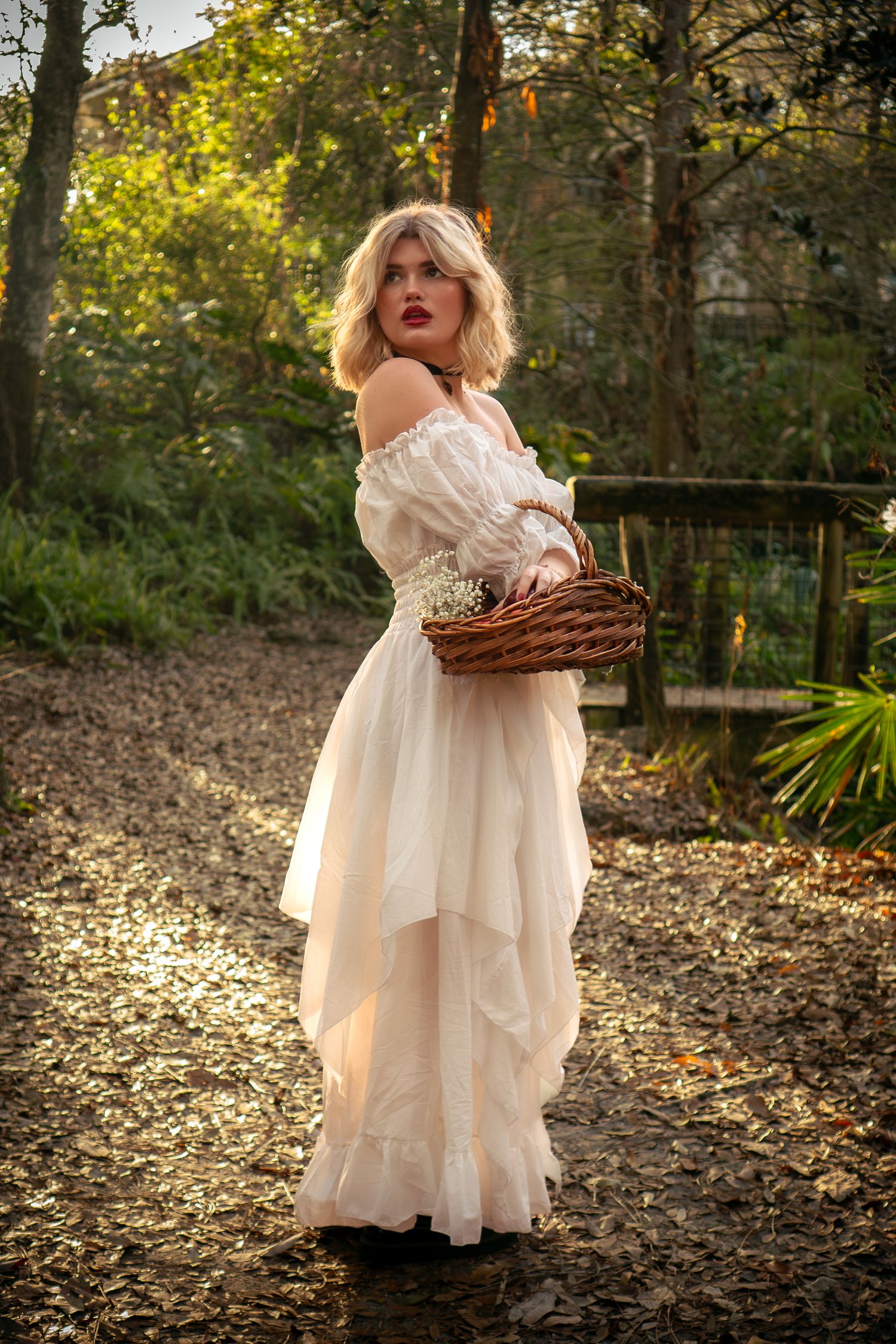 Blonde woman in white ruffled gown carrying wicker basket on a sunlit forest path.