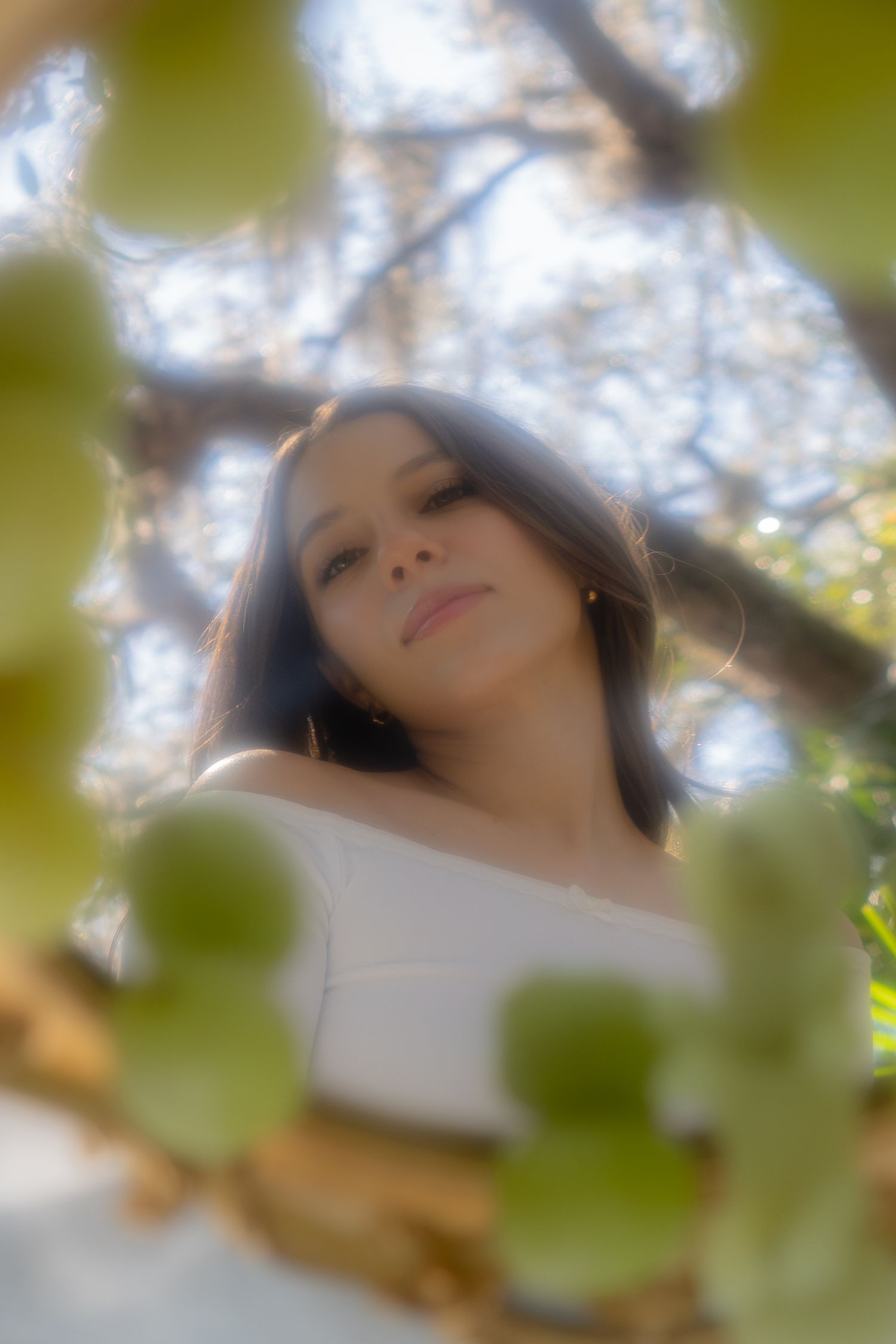 Brunette woman in white top photographed through green apple tree branches outdoors.