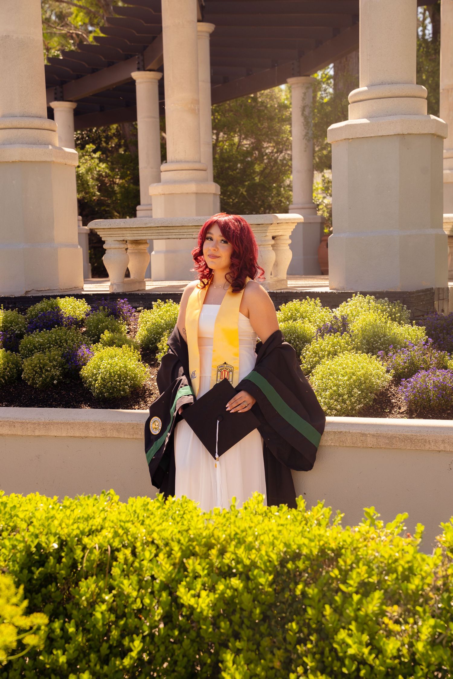 Red-haired graduate in white dress holds cap and stole beside garden columns in warm sunlight.