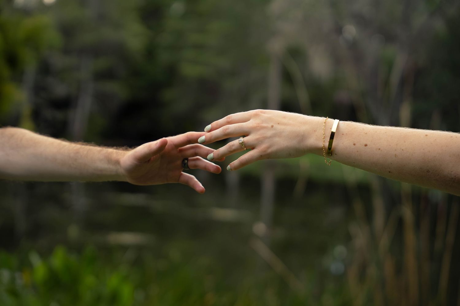 Two hands gently touching outdoors near a pond, with an engagement ring visible on the woman's finger.