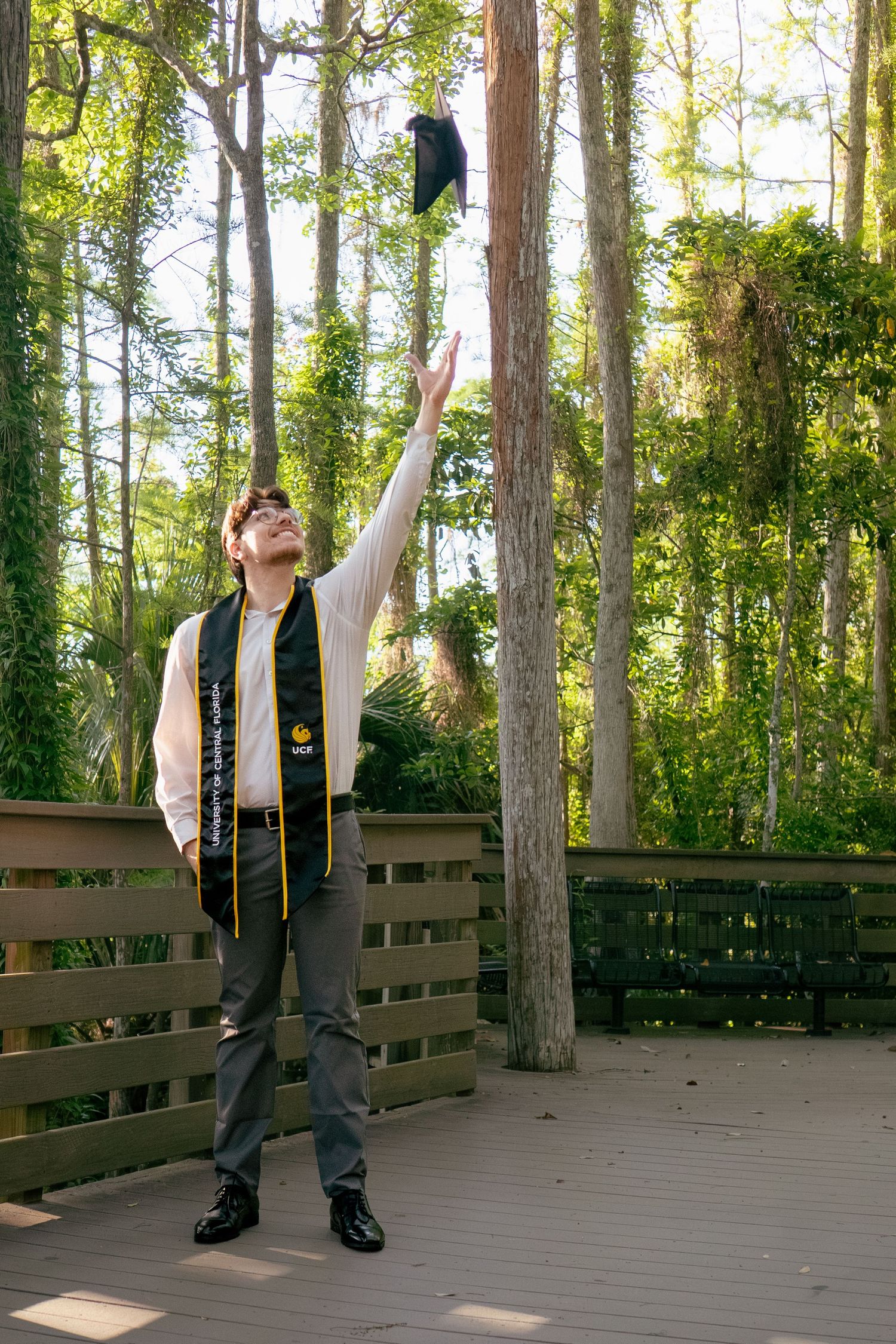 Male UCF graduate tossing black cap into the air on a wooden forest boardwalk.