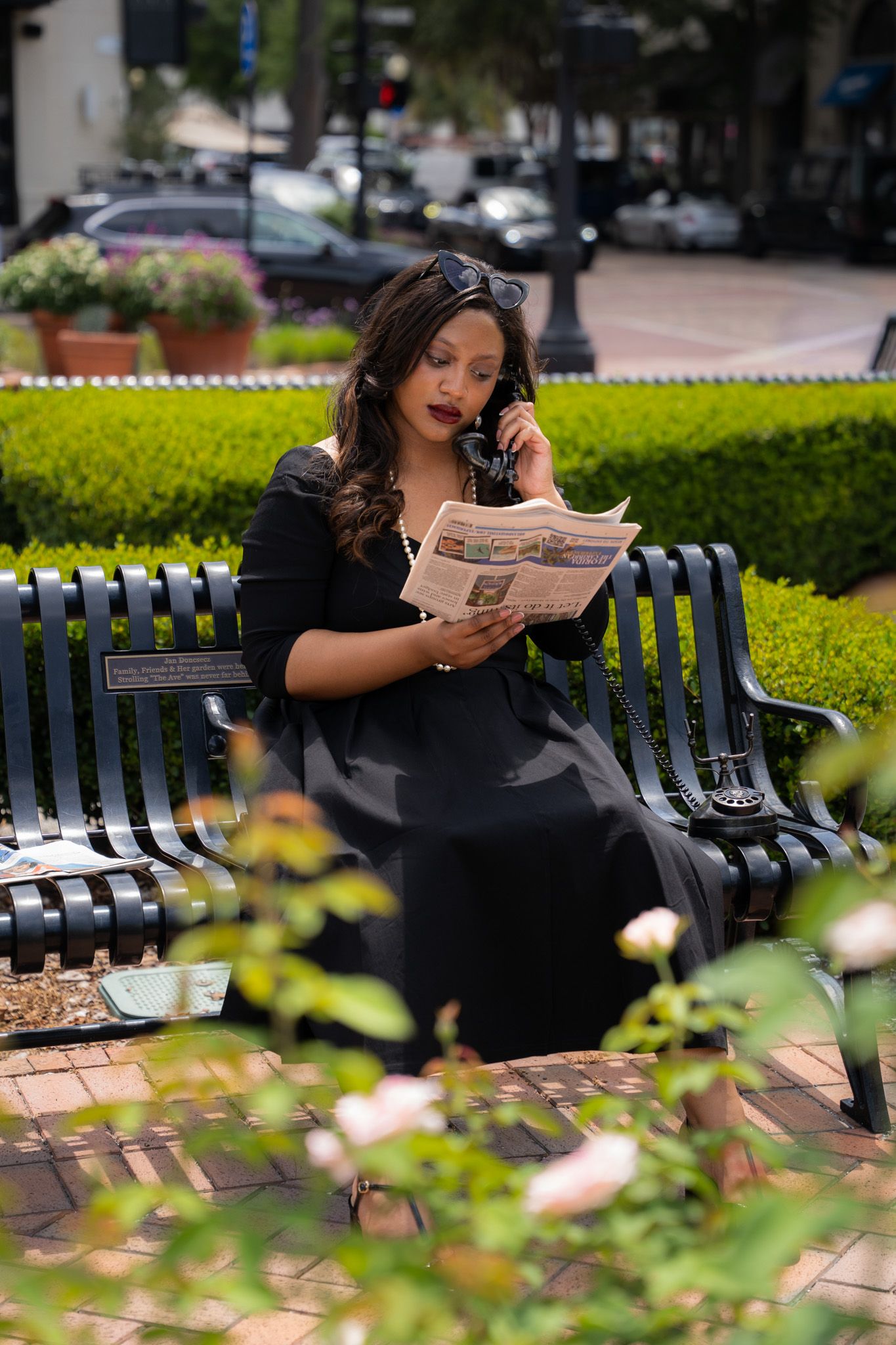Elegant woman in black dress sitting on a park bench reading a newspaper while on the phone.