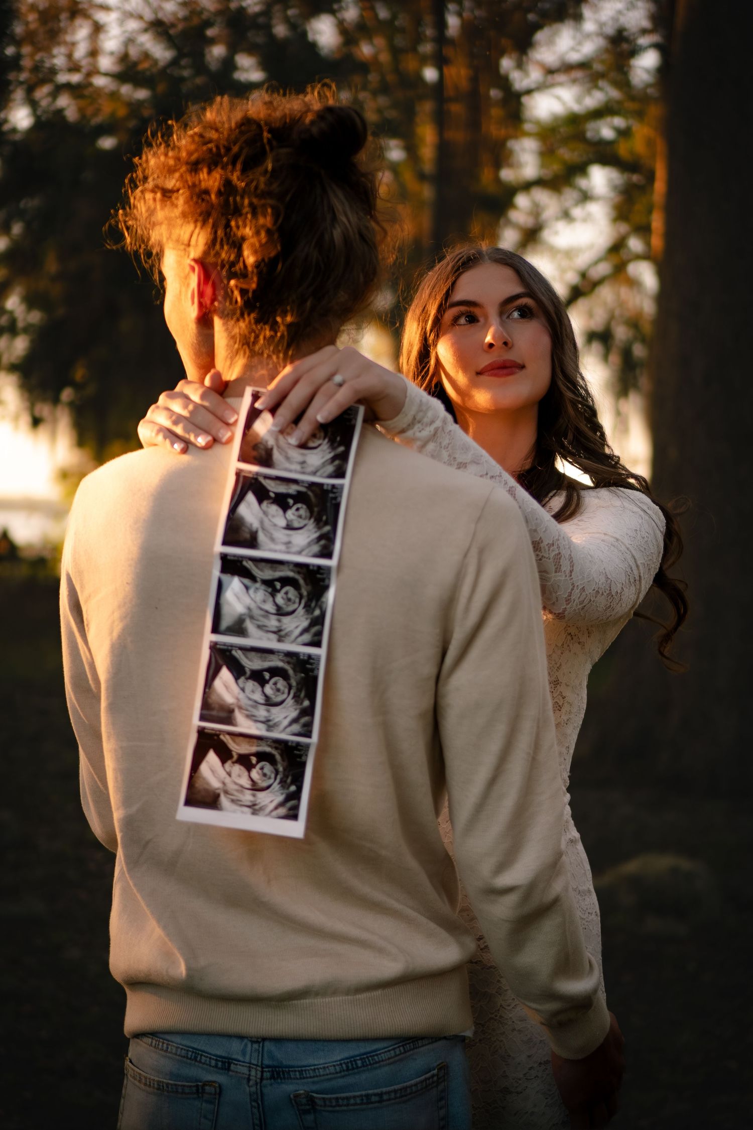 Pregnant woman in white lace dress embracing partner displaying ultrasound photos in sunset forest setting.