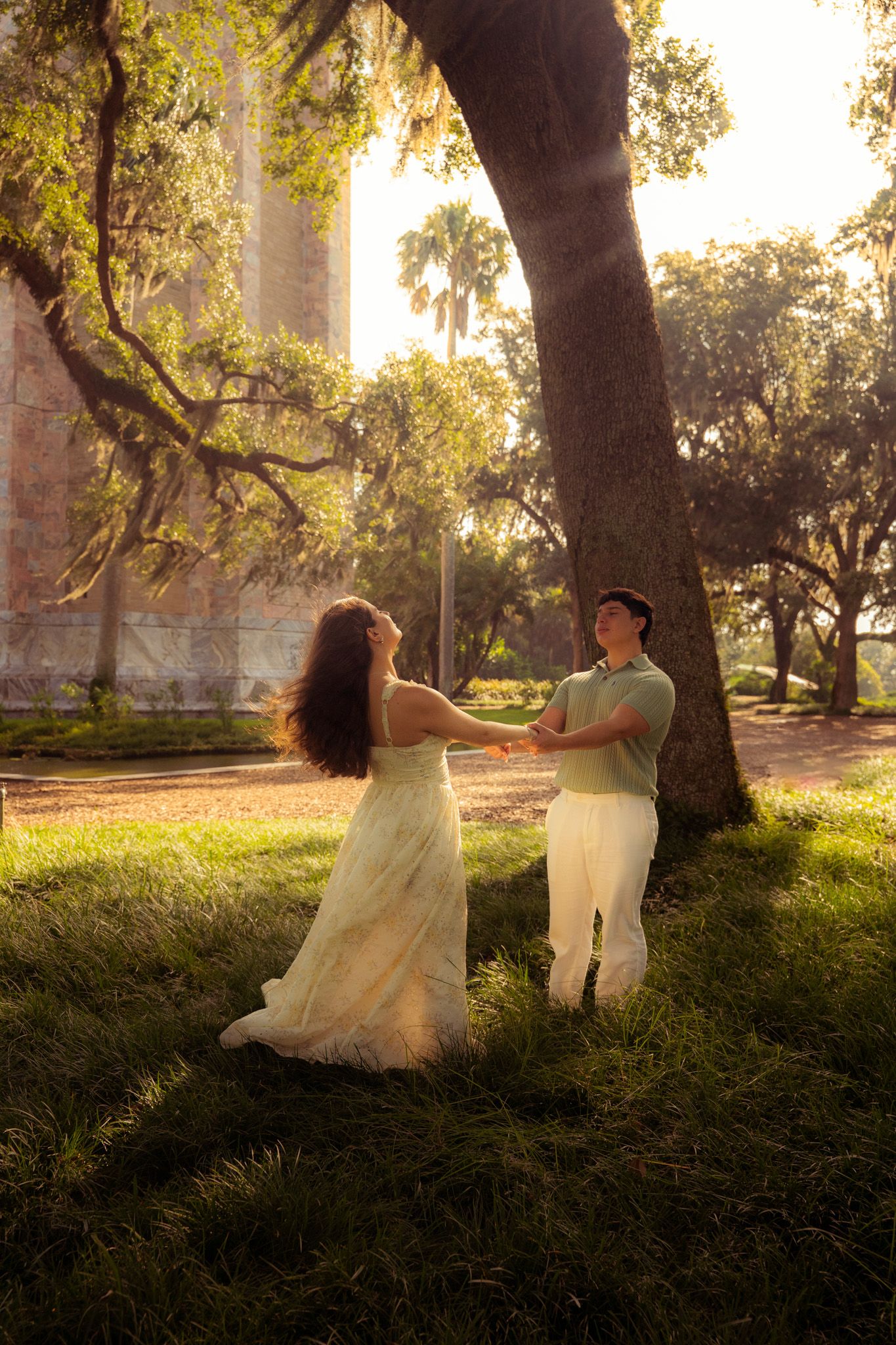 Couple dancing playfully hand in hand beneath a large oak tree near a historic stone building.