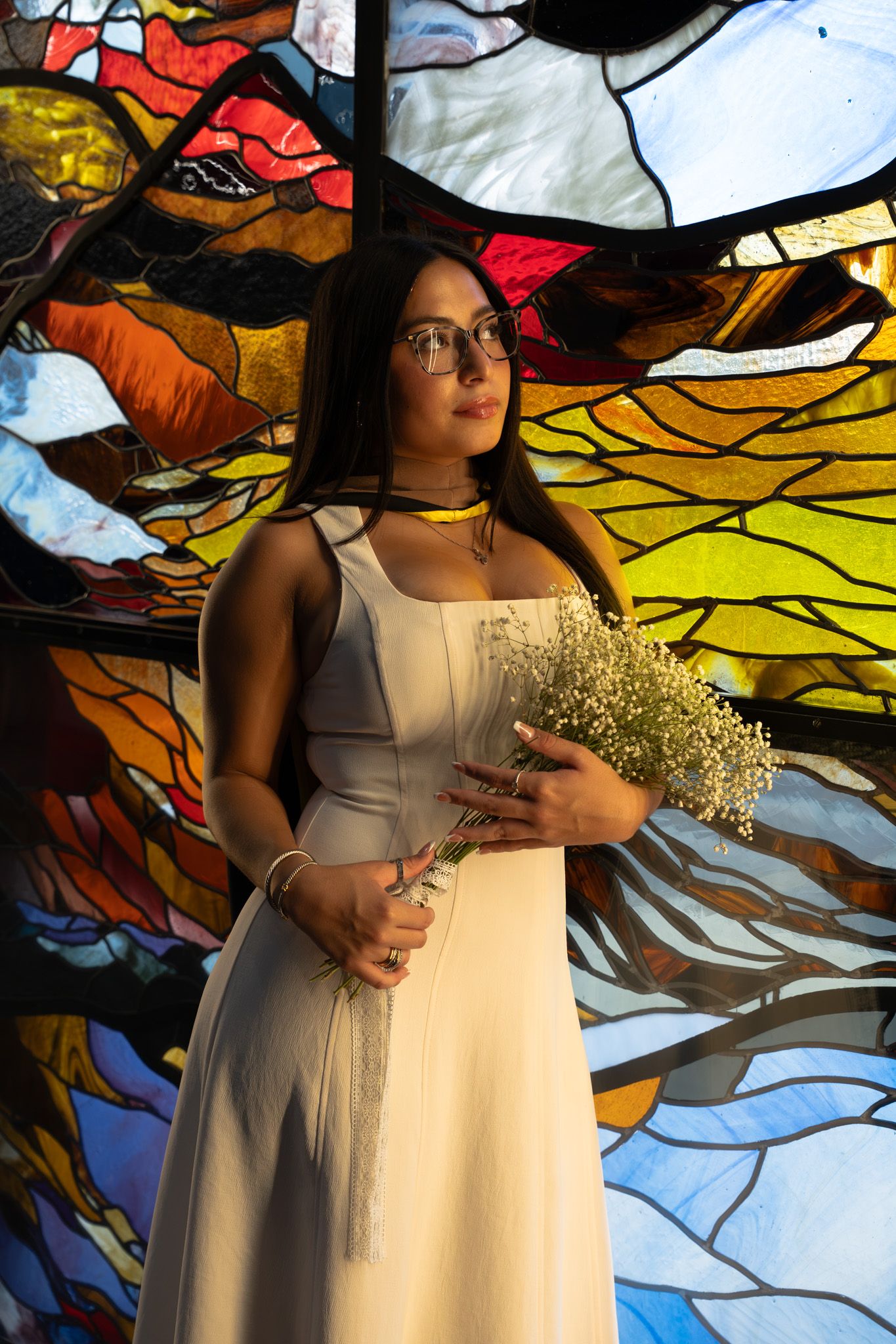 Young woman in white dress holding wildflowers poses before colorful stained glass window wearing glasses.