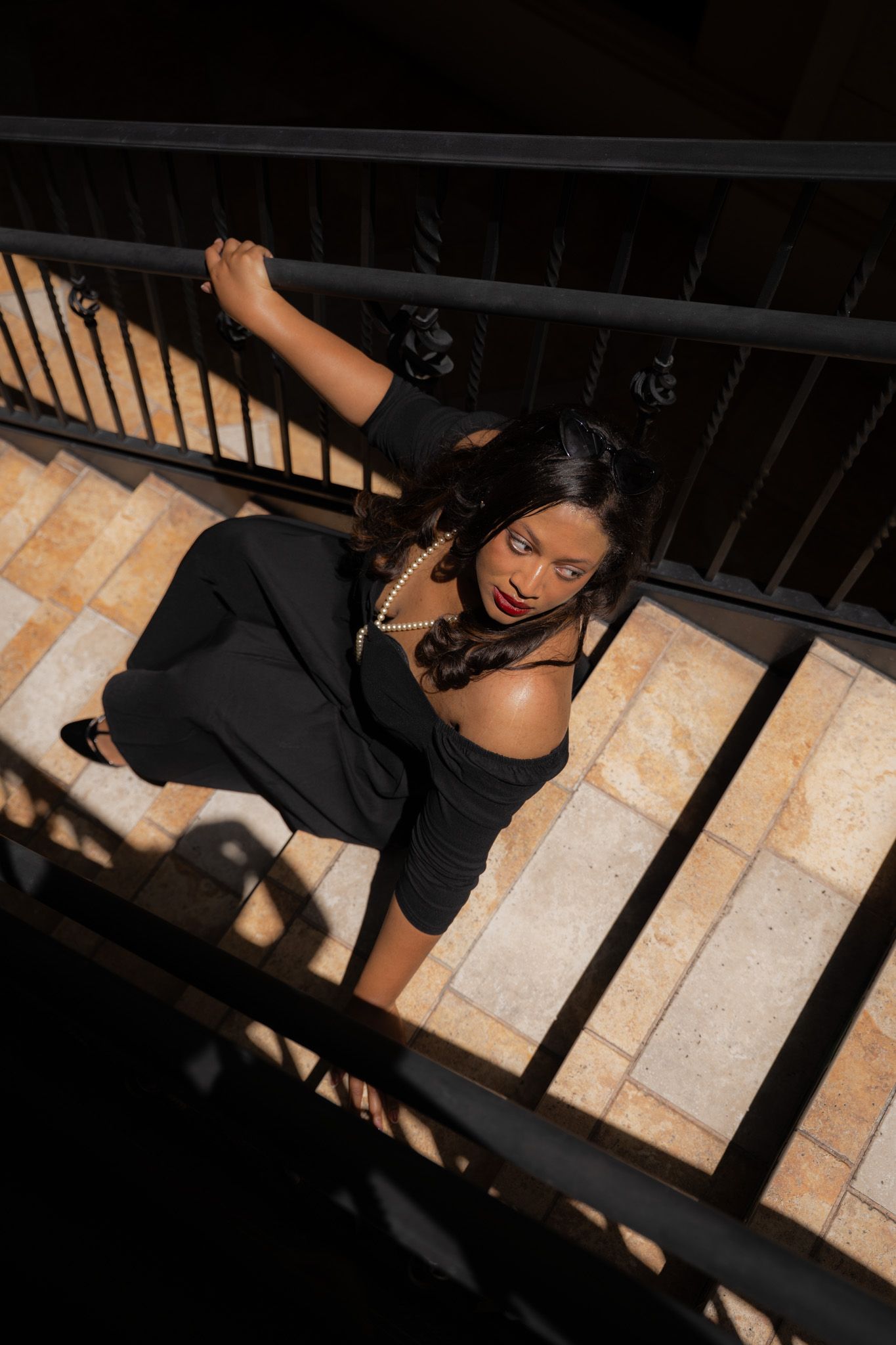 Elegant woman in black dress and pearls poses on sunlit stone staircase with iron railing.
