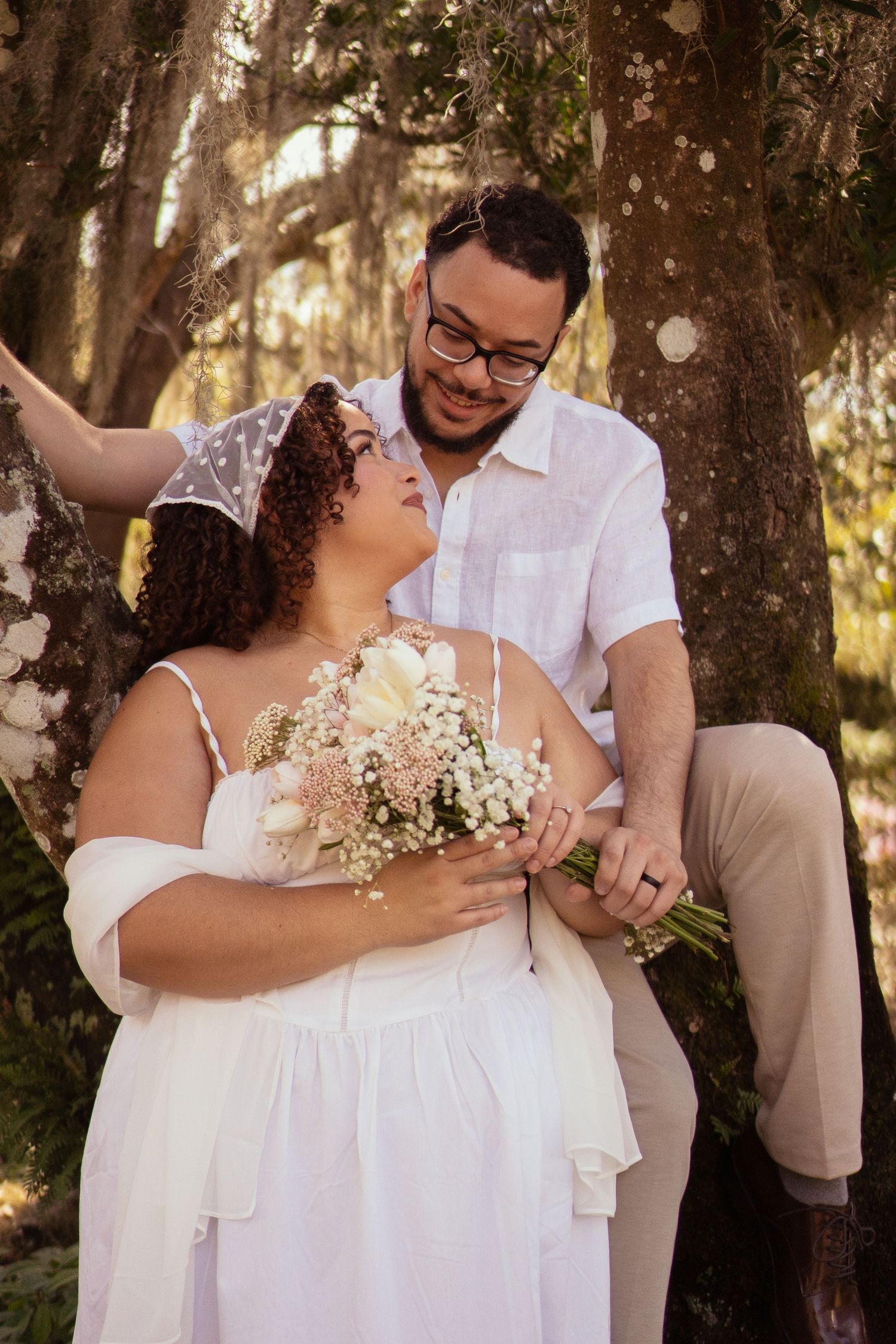 Smiling couple in white attire embracing by a mossy tree, bride holding a delicate floral bouquet.