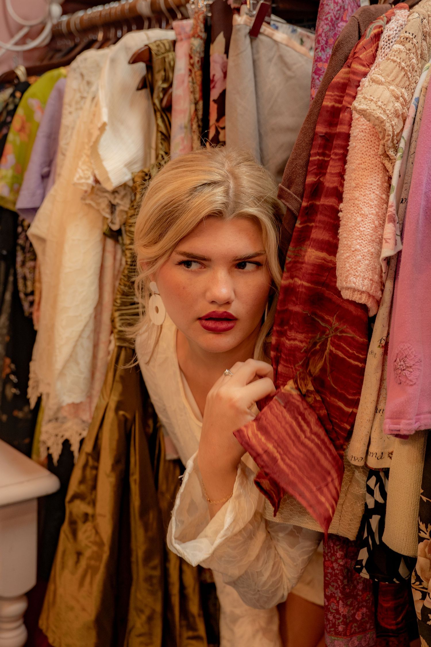 Blonde woman peering through colorful vintage clothing rack in a boutique shop.