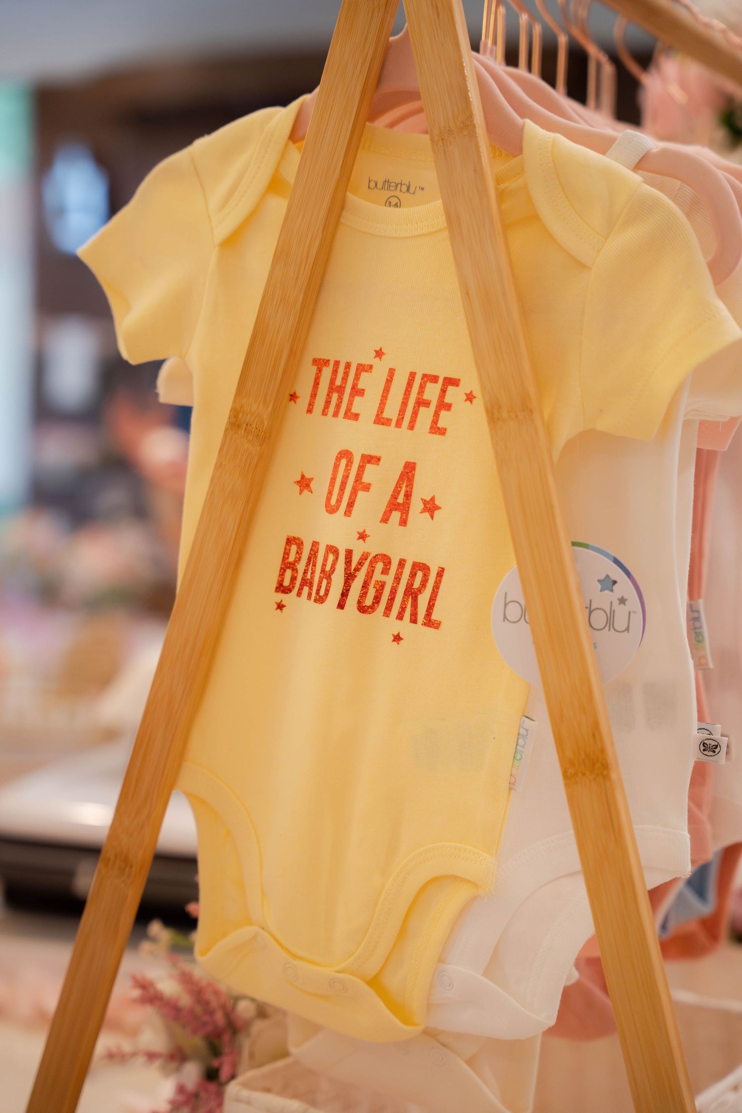 Yellow baby onesie with The Life of a Baby Girl text hanging on wooden display rack.