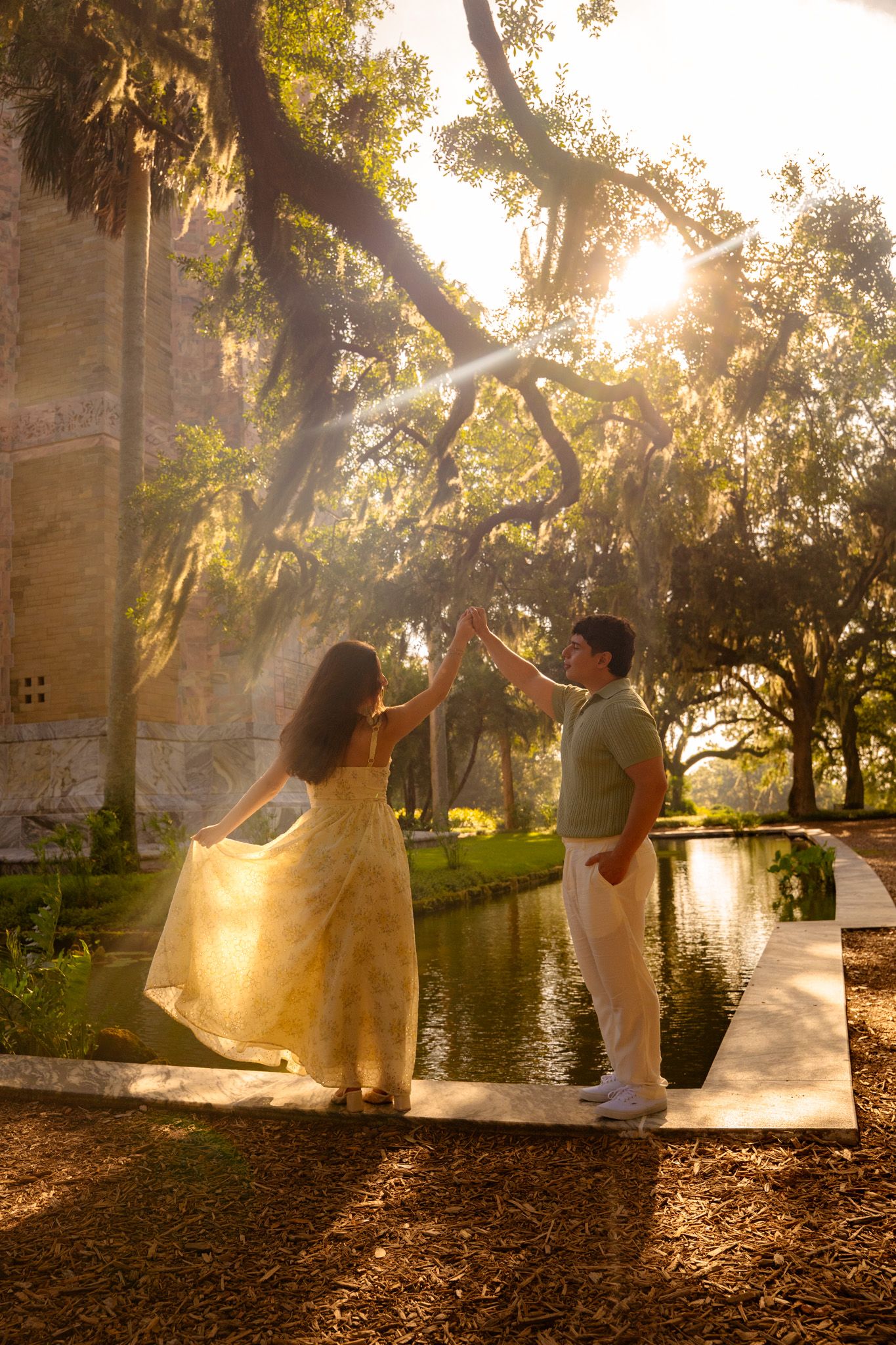 Couple dancing near sunlit pond with Spanish moss oak trees and historic building at sunset.