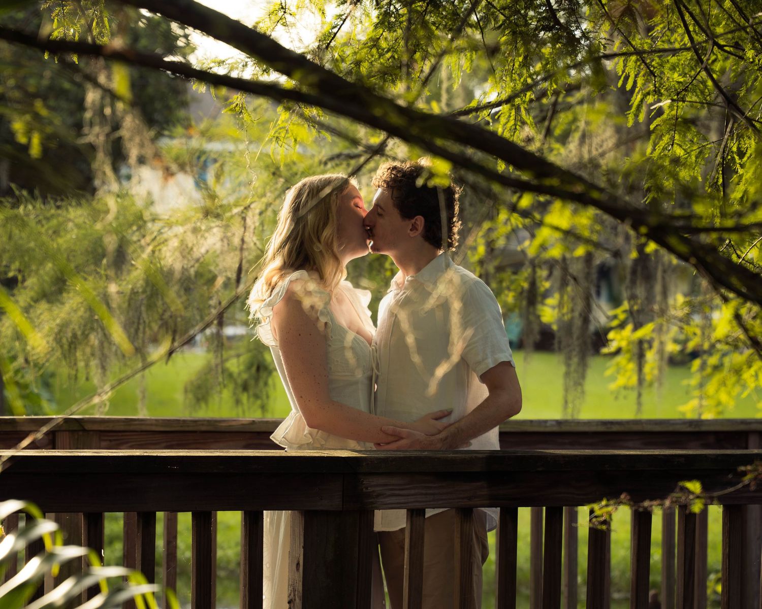 Couple sharing a romantic kiss on a wooden bridge surrounded by green trees.