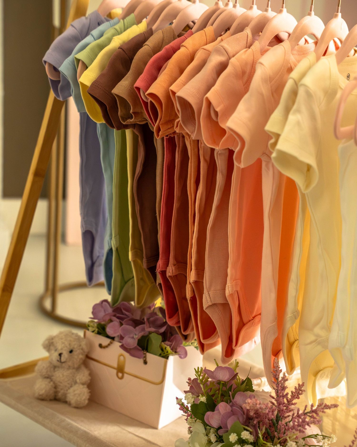 Colorful rainbow of baby onesies hanging on a wooden clothing rack with teddy bear decor.