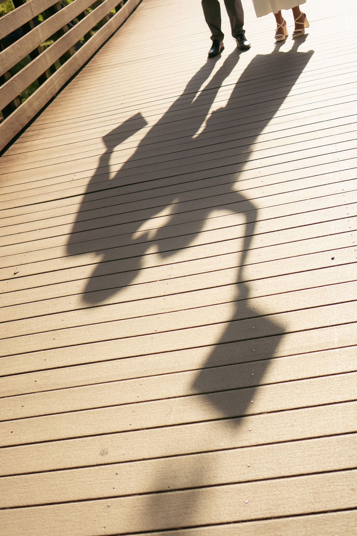 Shadows of two graduates holding caps cast on a sunny wooden boardwalk deck.